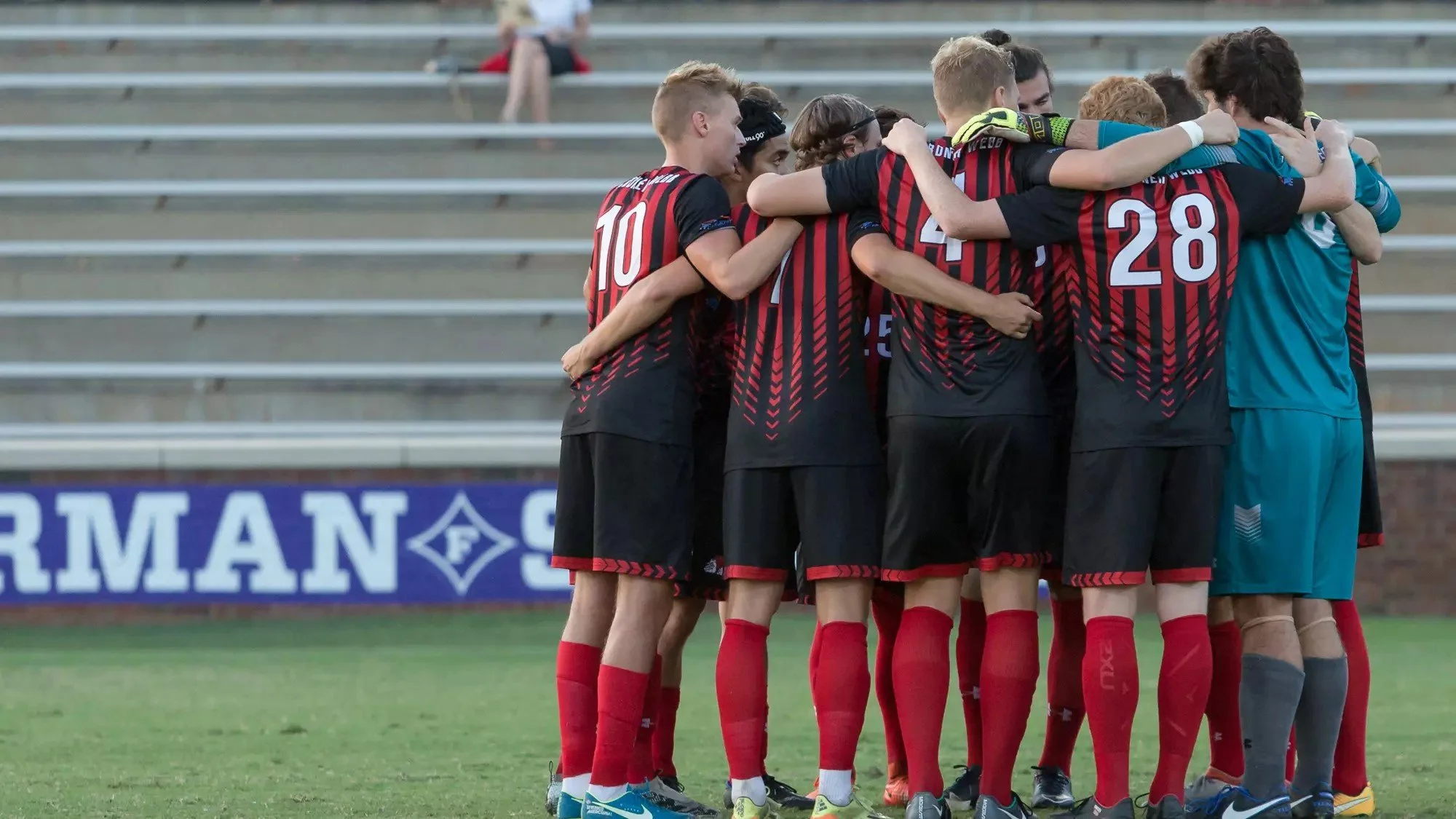 Division I college soccer team huddling together on the field before a match, with the goalkeeper wearing a teal jersey.