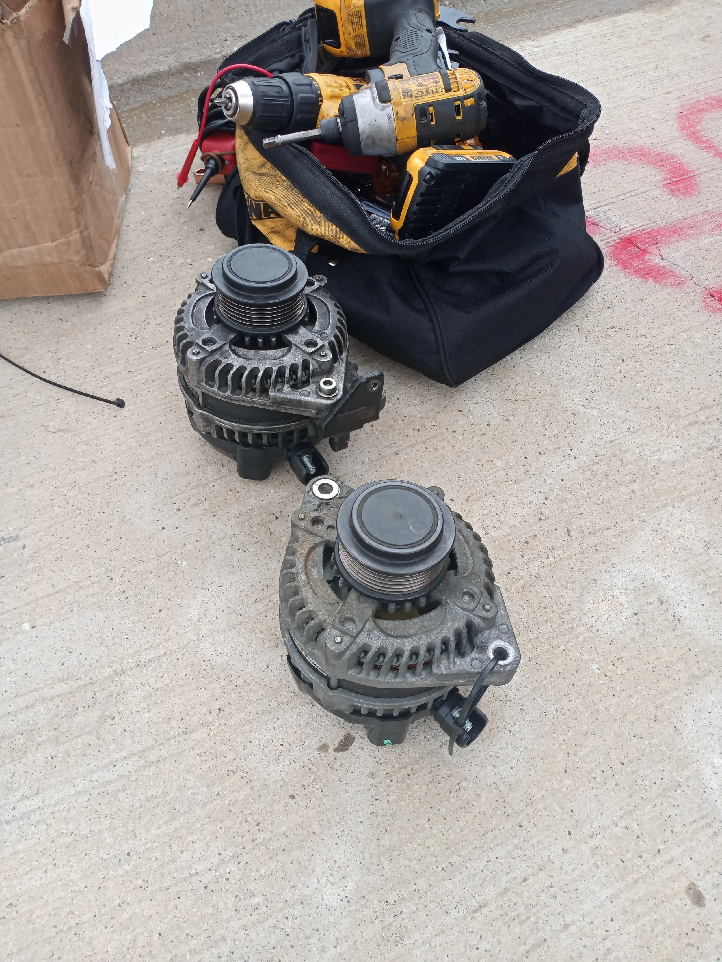 Two car alternators placed on a concrete ground outside, with tools and a black bag nearby.