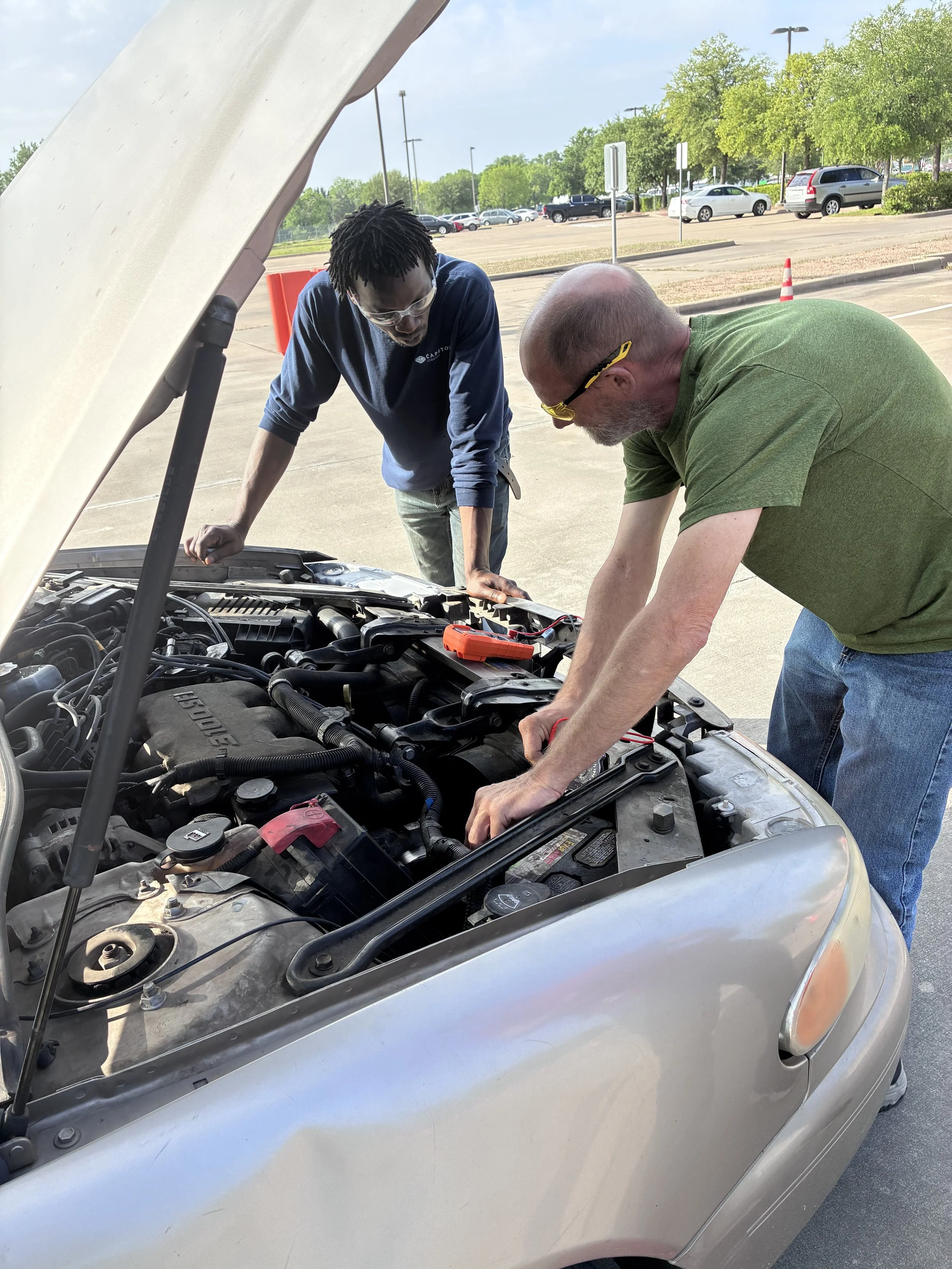 Two men inspecting a car's engine in a parking lot on a sunny day.