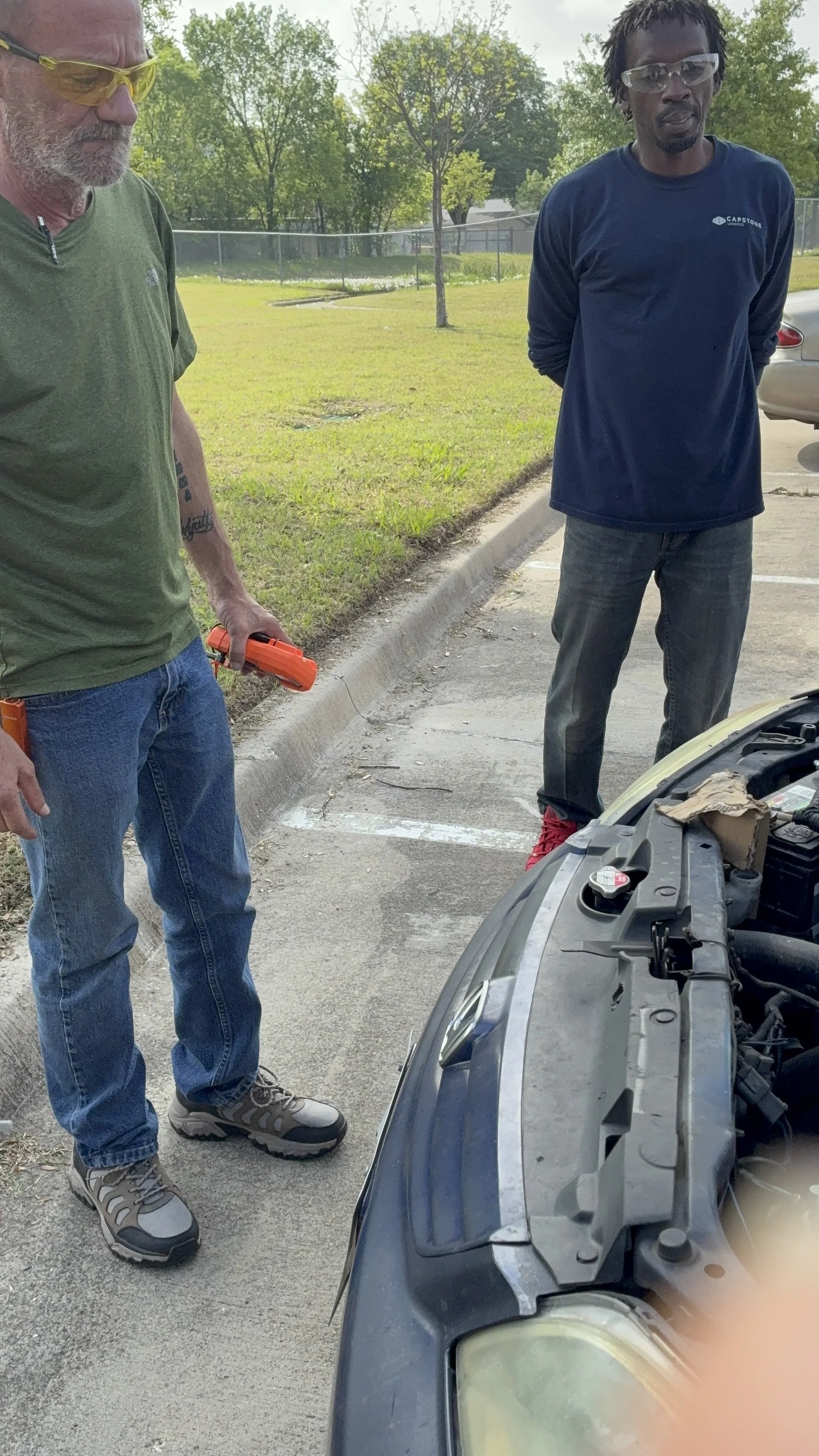 Two men inspecting a car engine in a parking lot, wearing safety glasses, one with gray hair in a green shirt holding an orange tool, the other with black dreadlocks in a navy shirt with hands behind back.
