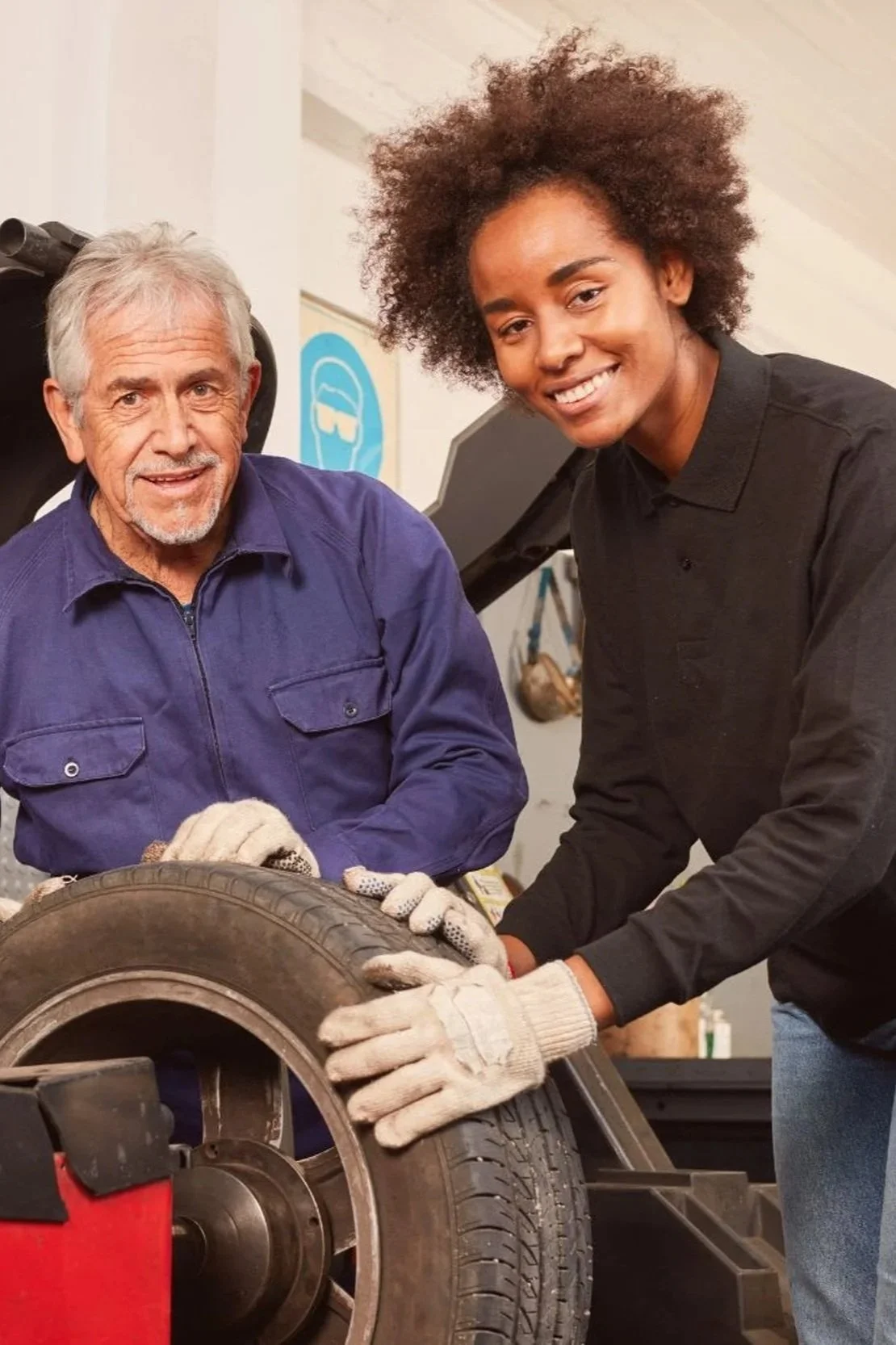 An elderly man and a young woman are working together on a tire at a workshop, both wearing gloves and smiling.