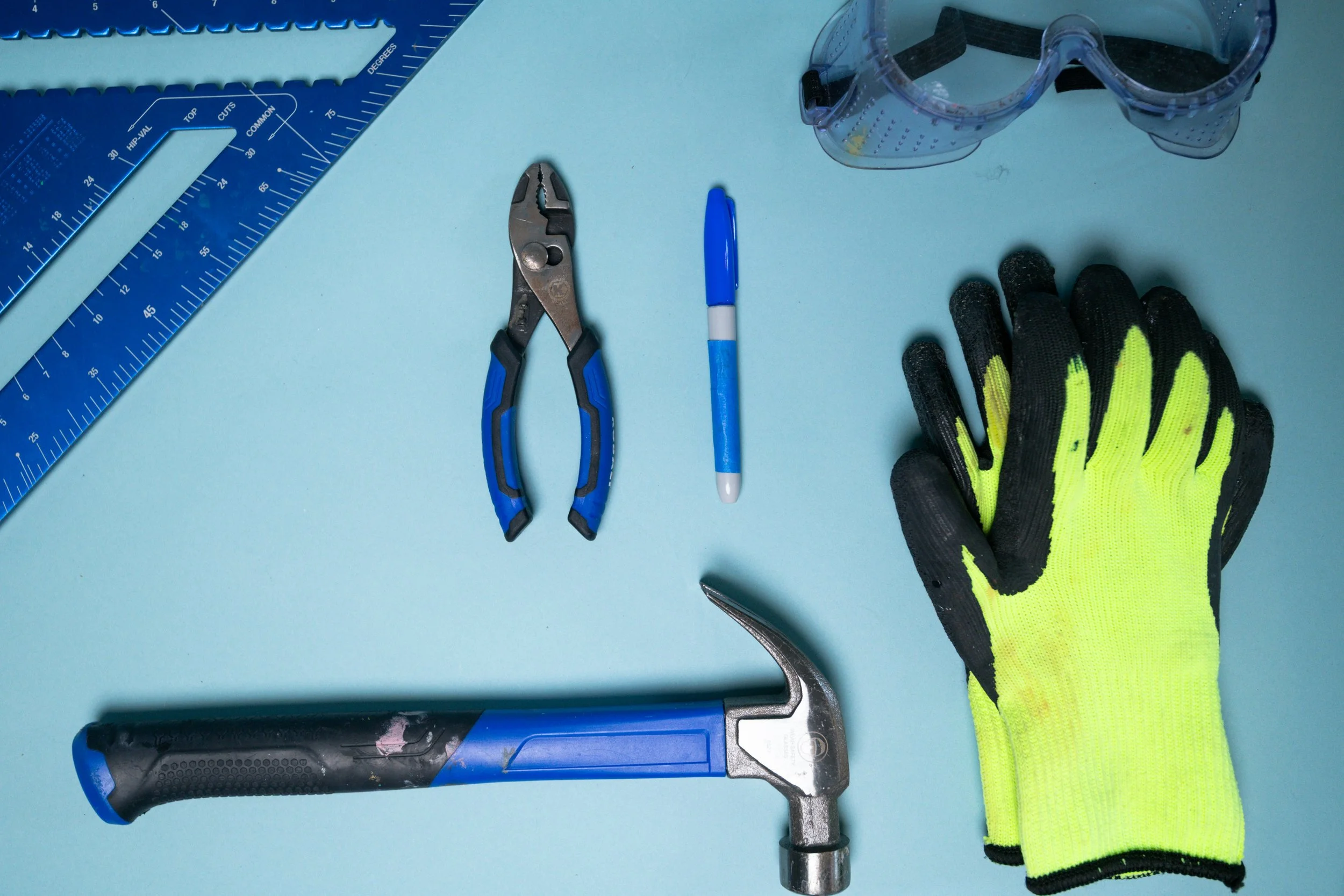 A light blue work surface with various tools including safety goggles, a pair of pliers, a blue marker, a cordless drill with a black and blue grip, a small portion of a blue ruler, and a pair of black and yellow work gloves.