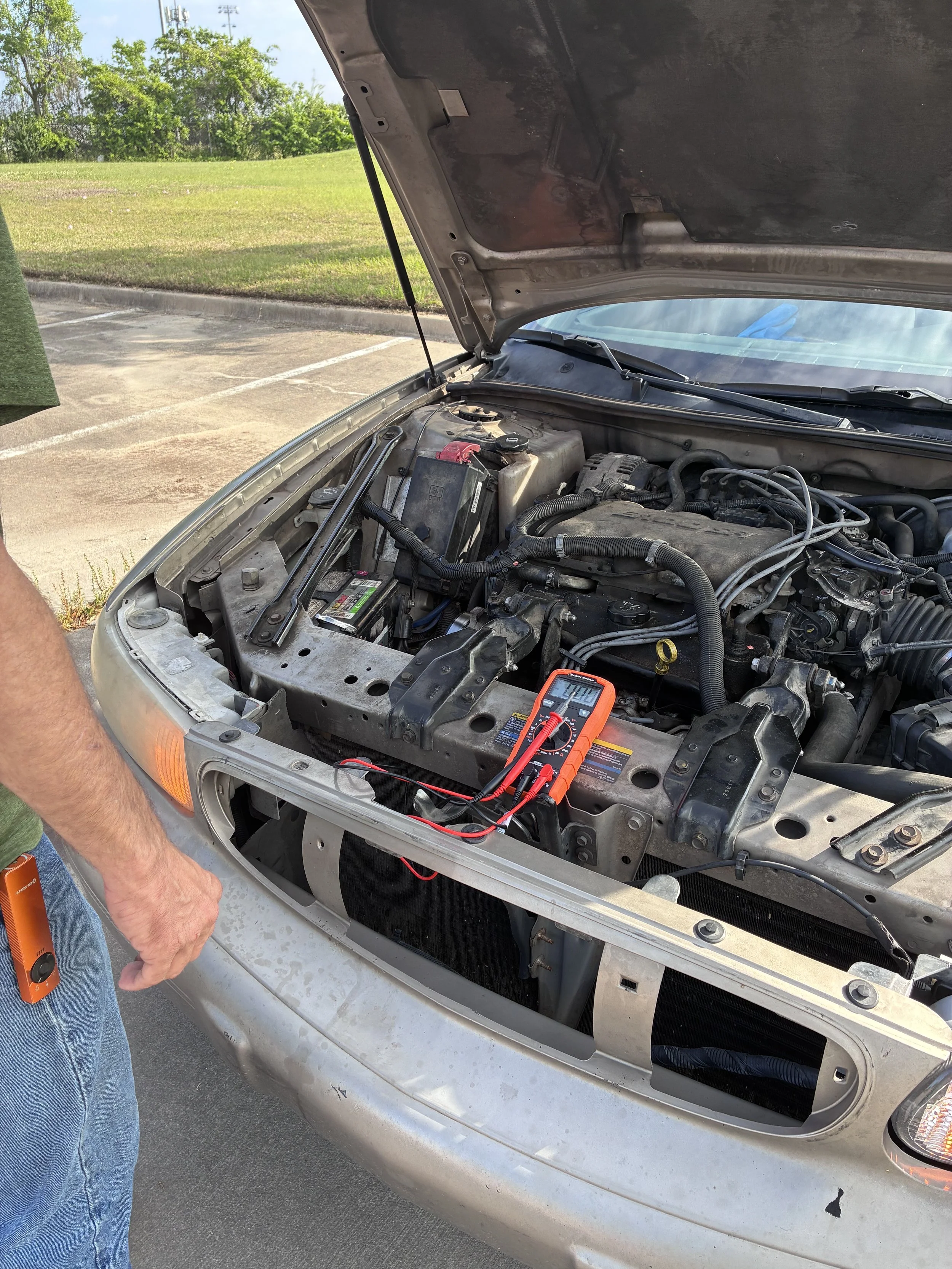 Open car hood showing engine bay with a person’s arm holding a multimeter attached to the battery in a parking lot.