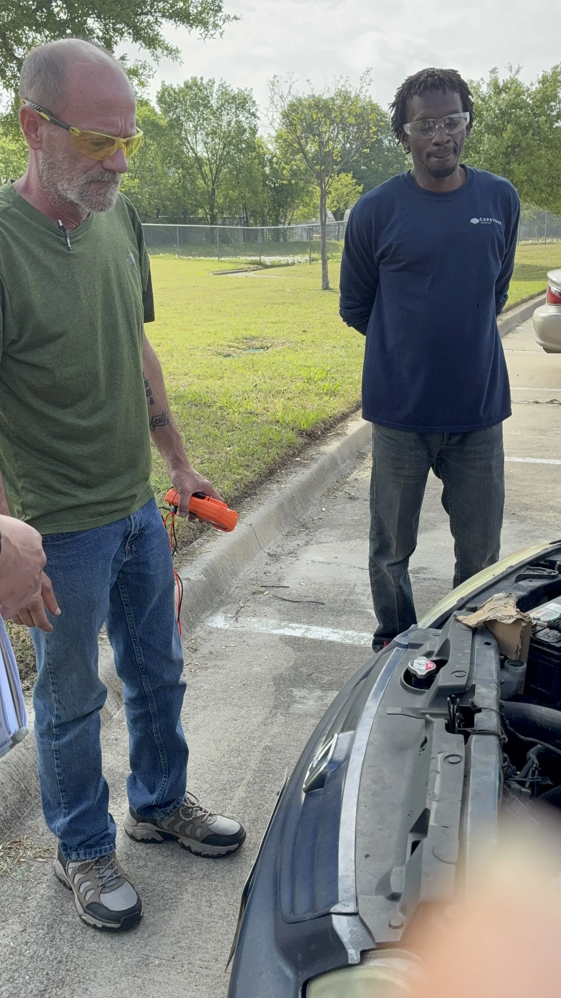 Two men standing on a parking lot inspecting a car engine, one holding a diagnostic tool, with green trees in the background.