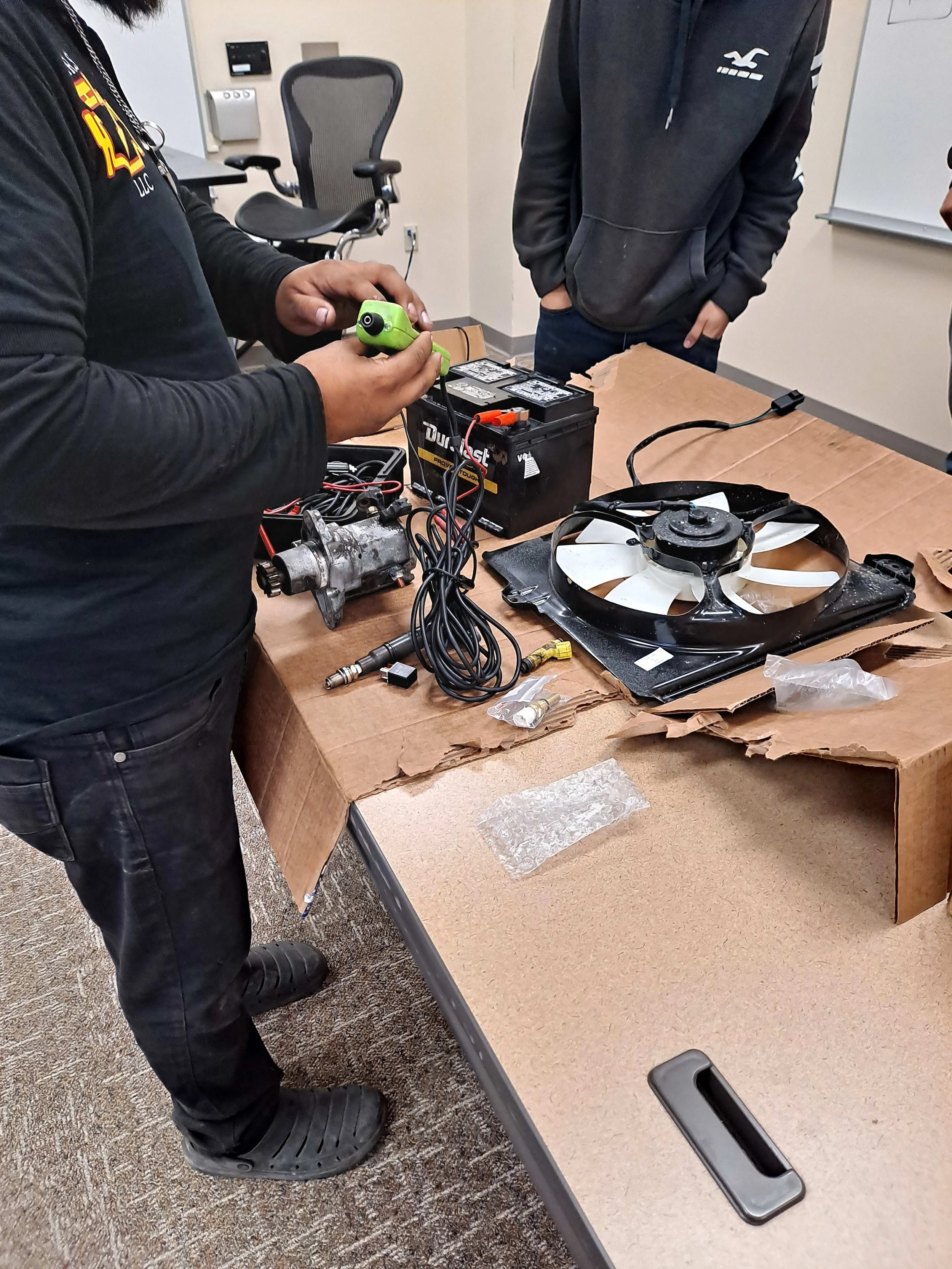 Individuals working with electronic equipment, including a battery, wires, and a fan, on a cardboard table in an indoor room.