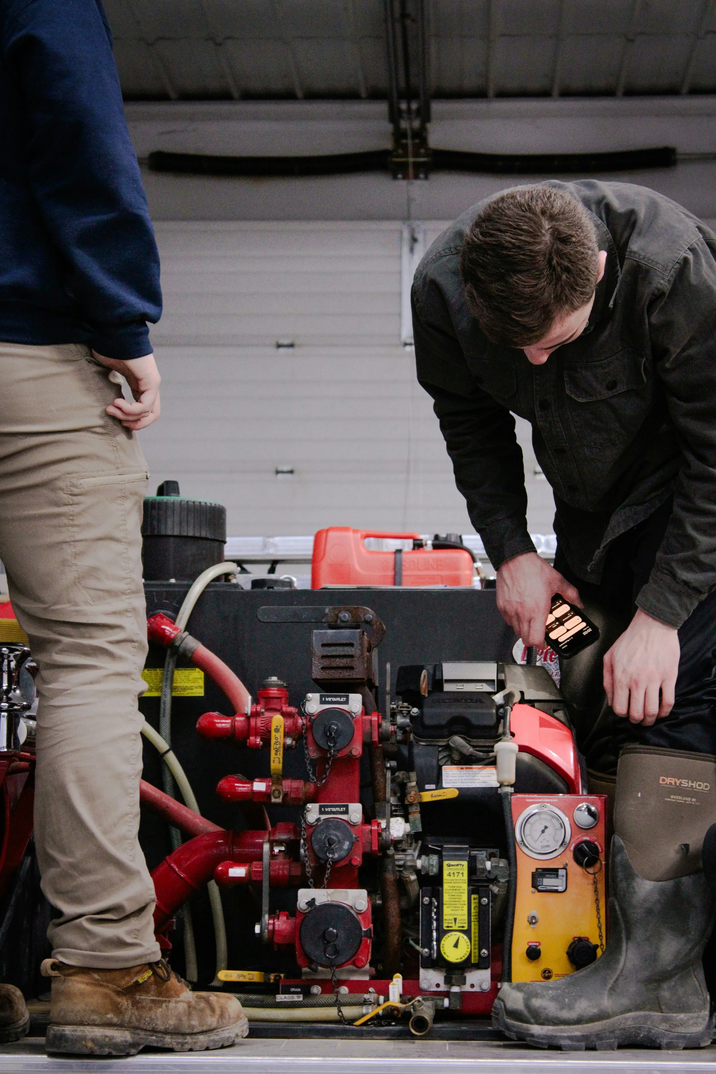 Two people working on a piece of machinery in a garage or industrial setting. One person is wearing tan work boots and pants, and the other is wearing dark work boots and pants. The machinery is red and orange with various pipes, gauges, and control panels.