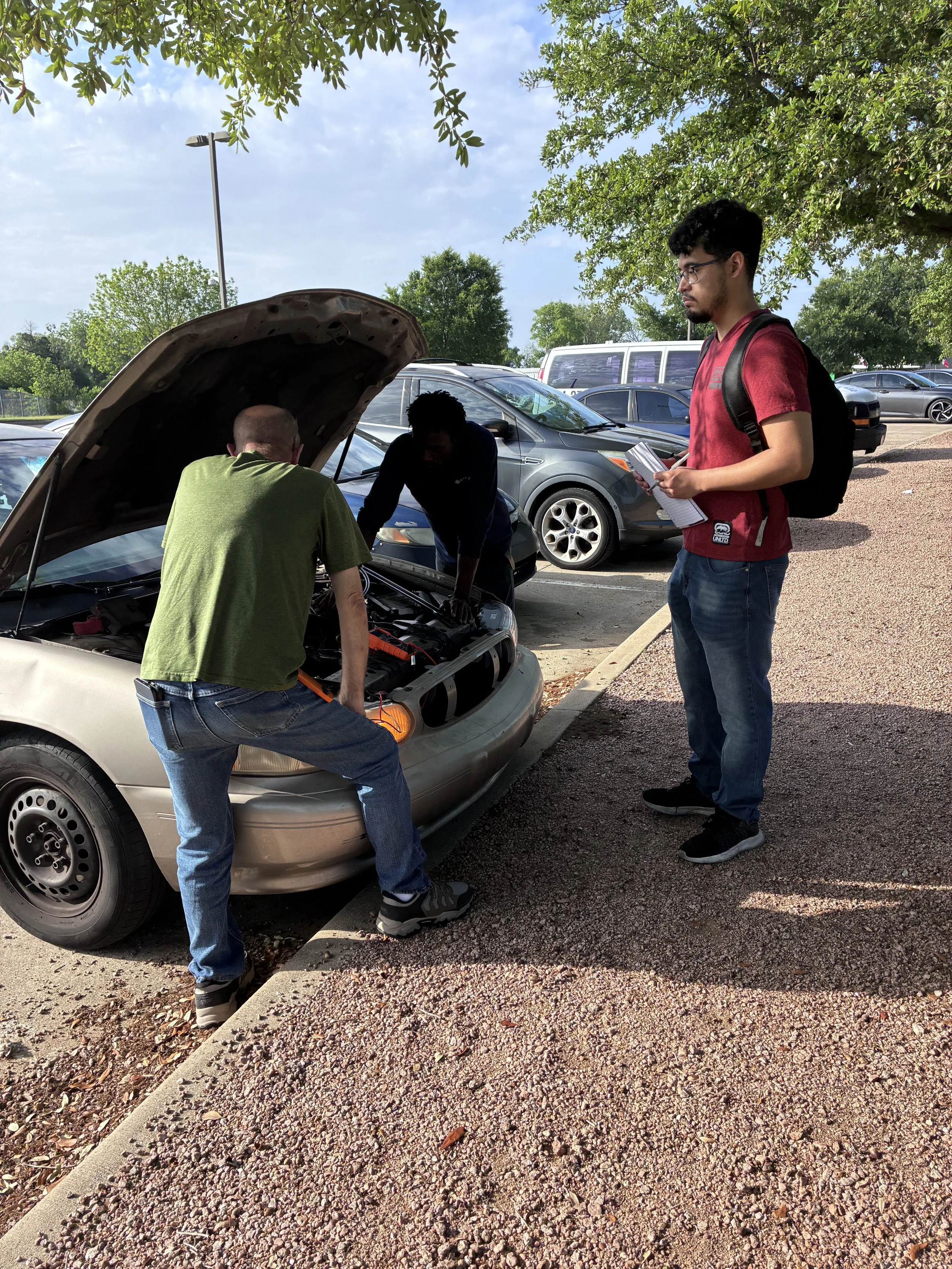 A young man with a backpack and glasses standing outside in a parking lot, observing two men working on a car with its hood open.
