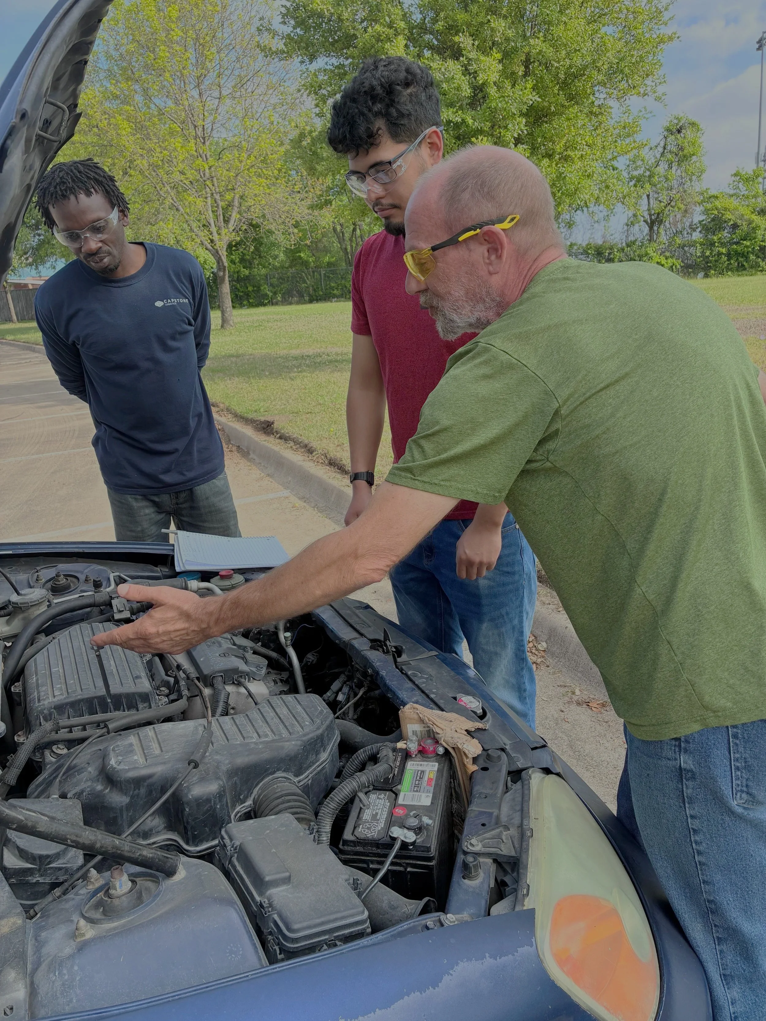 Three men with safety glasses inspecting a car engine outdoors on a sunny day.