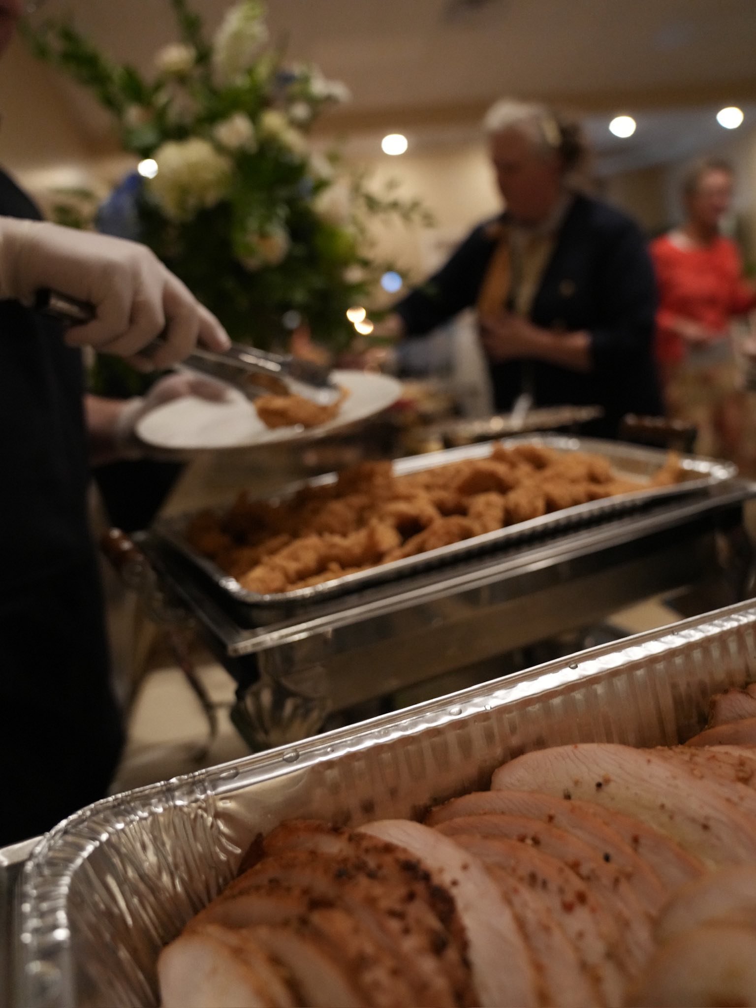 Buffet with trays of sliced chicken and fried chicken, with people in the background serving themselves at a holiday meal.
