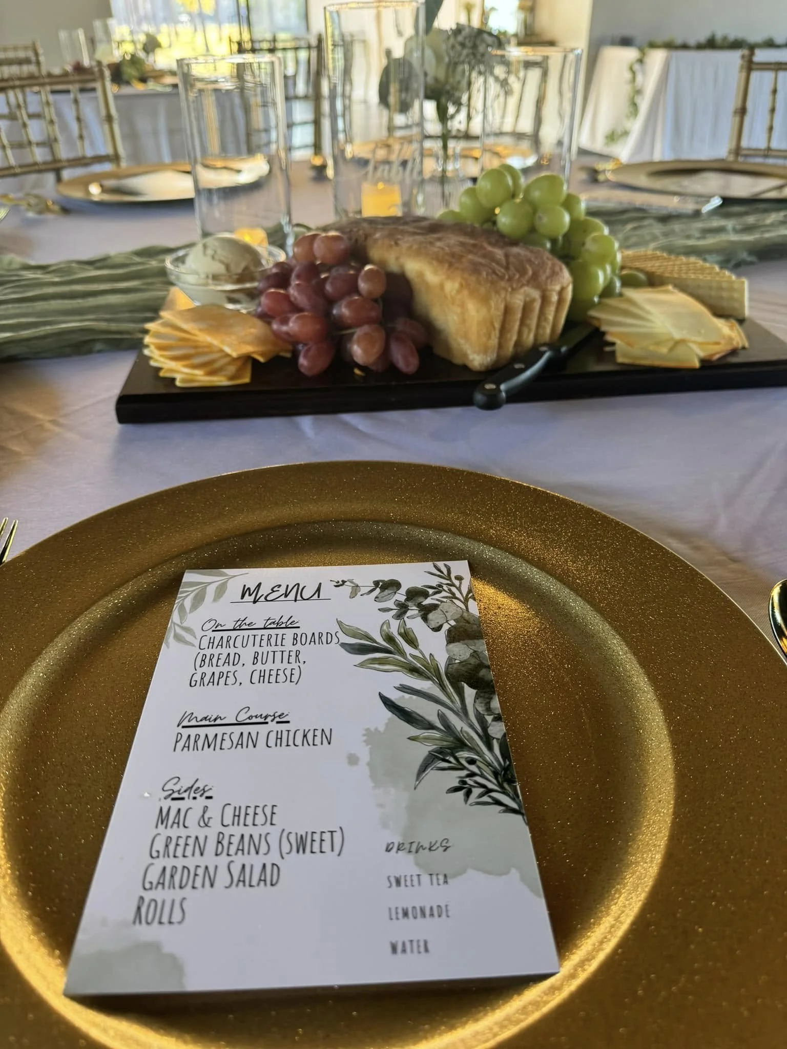 A table setting with a gold plate holding a menu and a charcuterie board in the background featuring bread, cheese, grapes, and crackers.