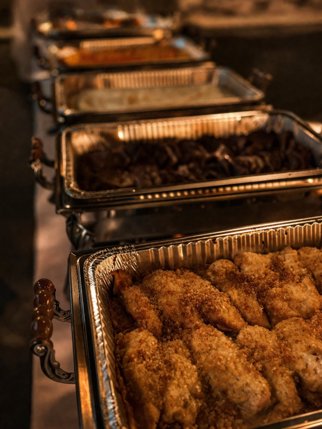 Chafing dishes with fried chicken and other cooked meats at a buffet or catering setup.
