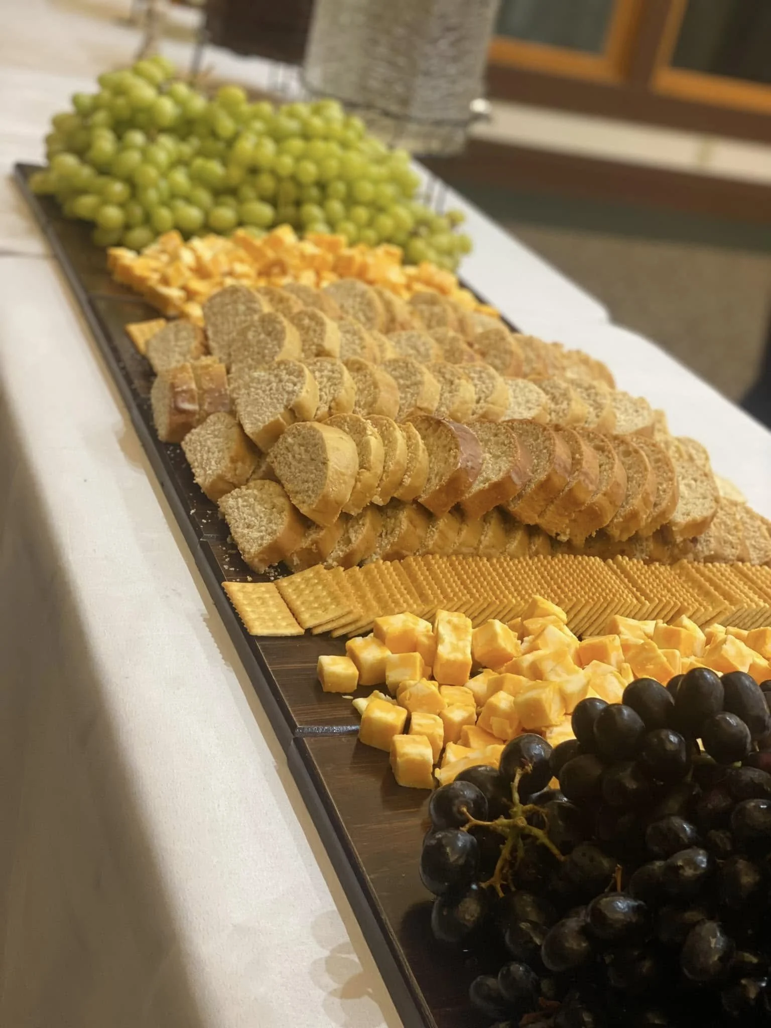 A long serving platter with green grapes, shredded cheese, sliced sausage, assorted crackers, cubed cheese, and black grapes.