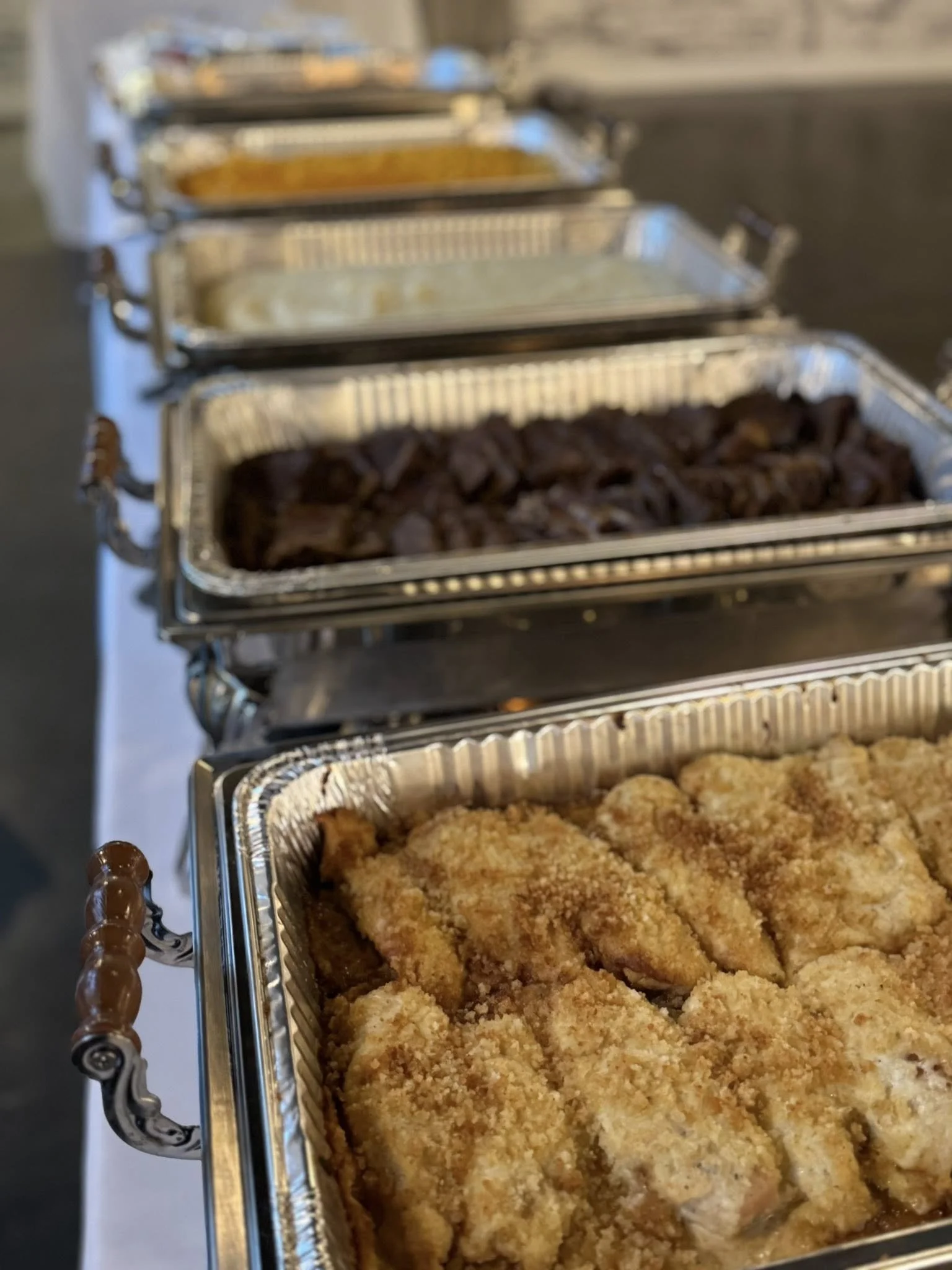 A row of chafing dishes with different foods, including breaded fried chicken in the foreground, cooked meat in the middle, and other dishes further back, set on a white tablecloth.