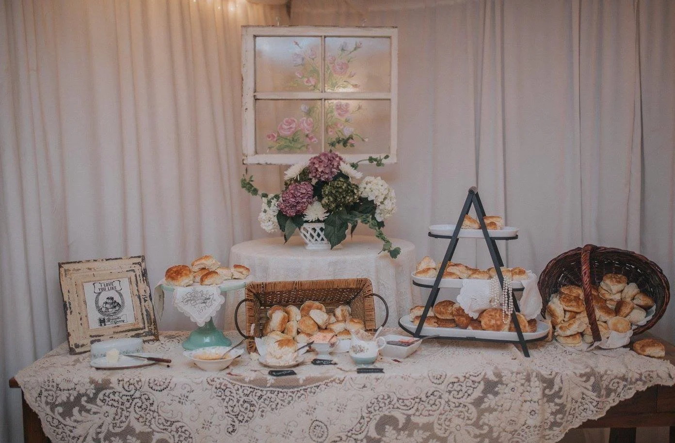A table decorated with a lace tablecloth, holding various pastries and buns, with a framed picture, a basket, and a tiered stand filled with baked goods. Behind the table, there is a floral arrangement on a small round table and a window with pastel-