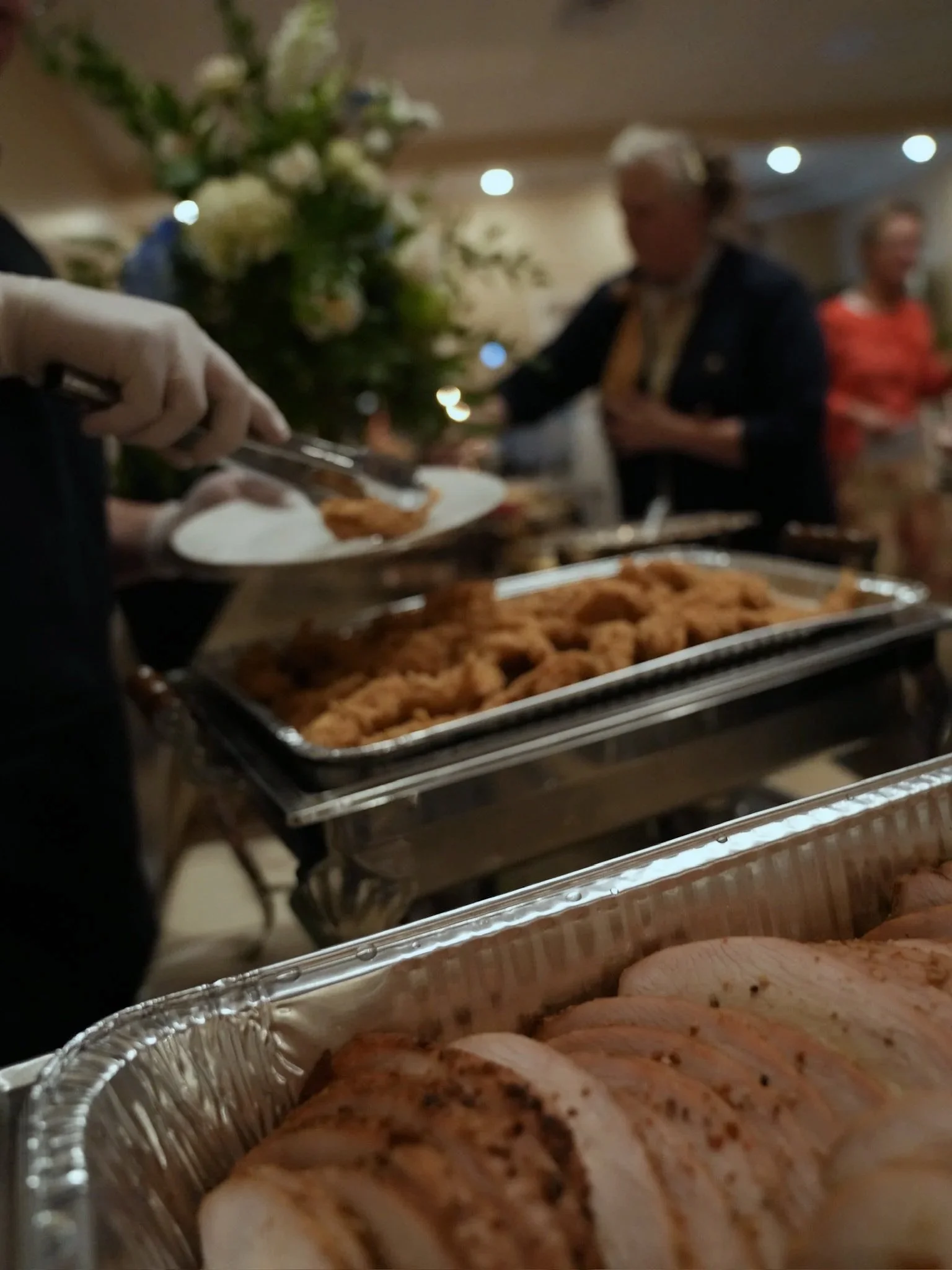 Buffet table with sliced cooked poultry in an aluminum tray, fried chicken in another tray, and a person serving food with tongs, with a large flower arrangement and guests in the background.