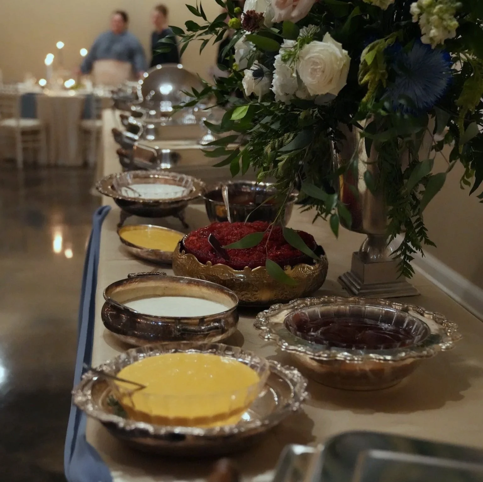 A table set with various bowls of sauces or condiments, a vase of white and blue flowers, and a blurred background of people in a dining or event setting.