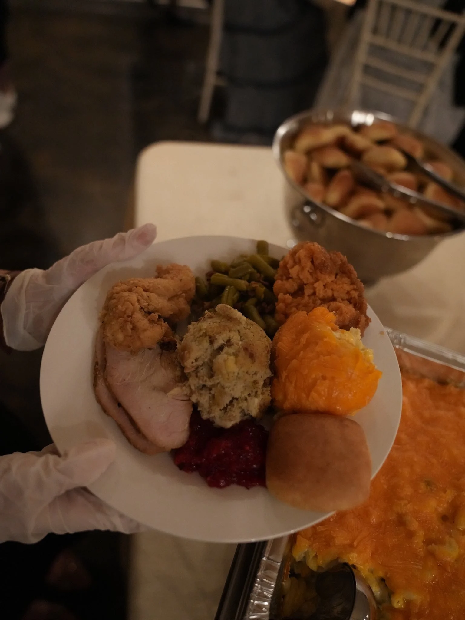 A person wearing gloves holding a white plate with fried chicken, green beans, stuffing, ham, sweet potato casserole, meatloaf, and cranberry sauce. In the background, there is a bowl of baked macaroni and cheese.