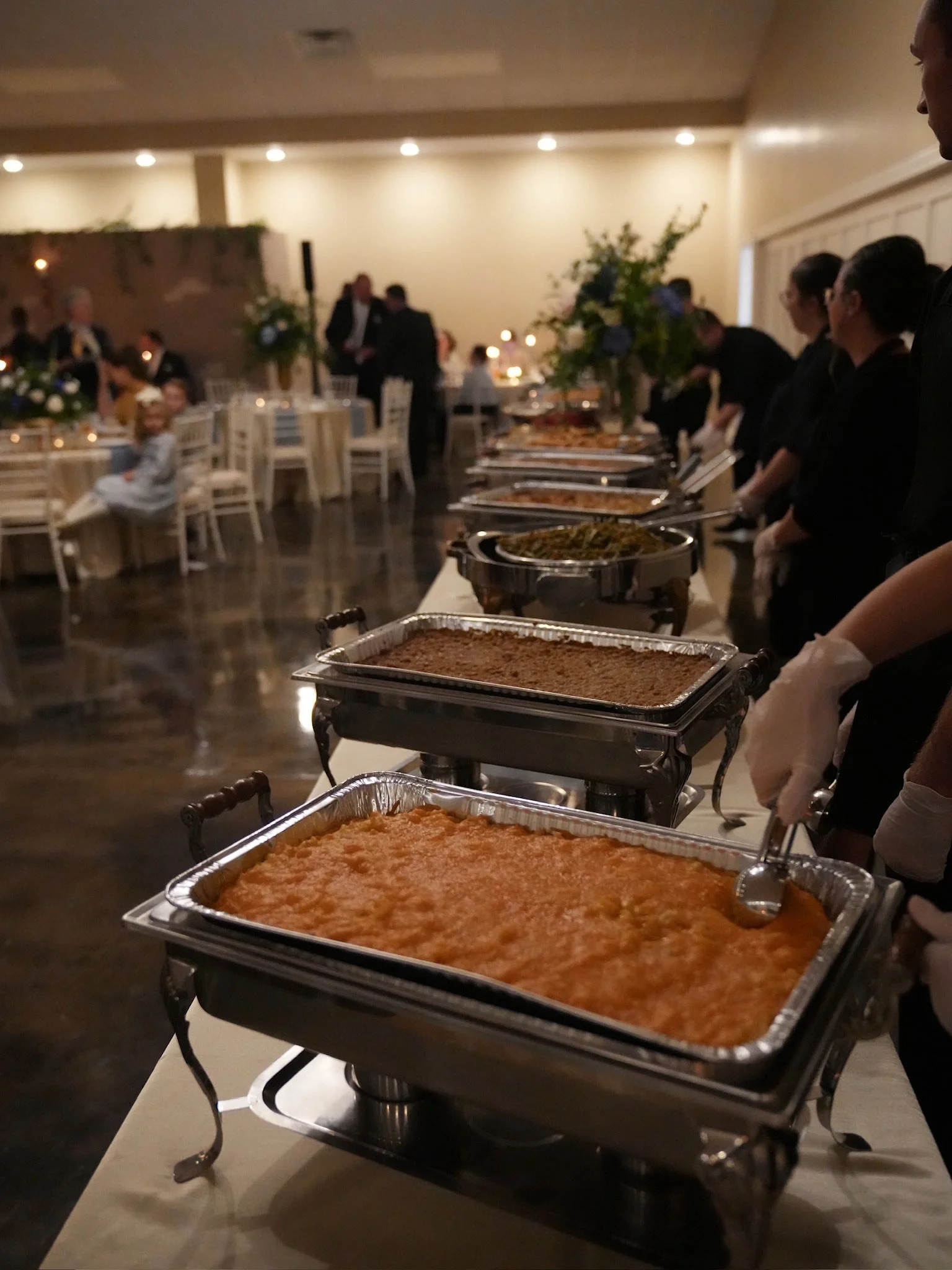Buffet table with chafing dishes filled with various hot dishes at a formal event in a dimly lit room with decorated tables and guests in the background.