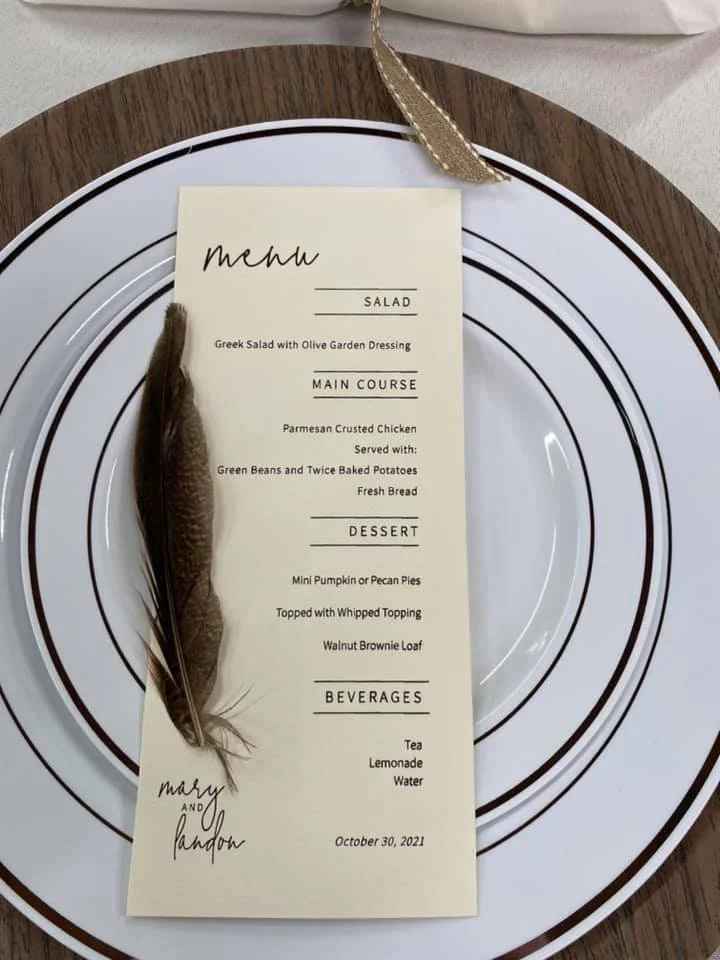 Dinner menu on a card placed on a decorative plate, with a feather and a ribbon. The menu includes Greek salad with olive garden dressing, Parmesan crusted chicken with green beans and baked potatoes, mini pumpkin or pecan pies topped with whipped to