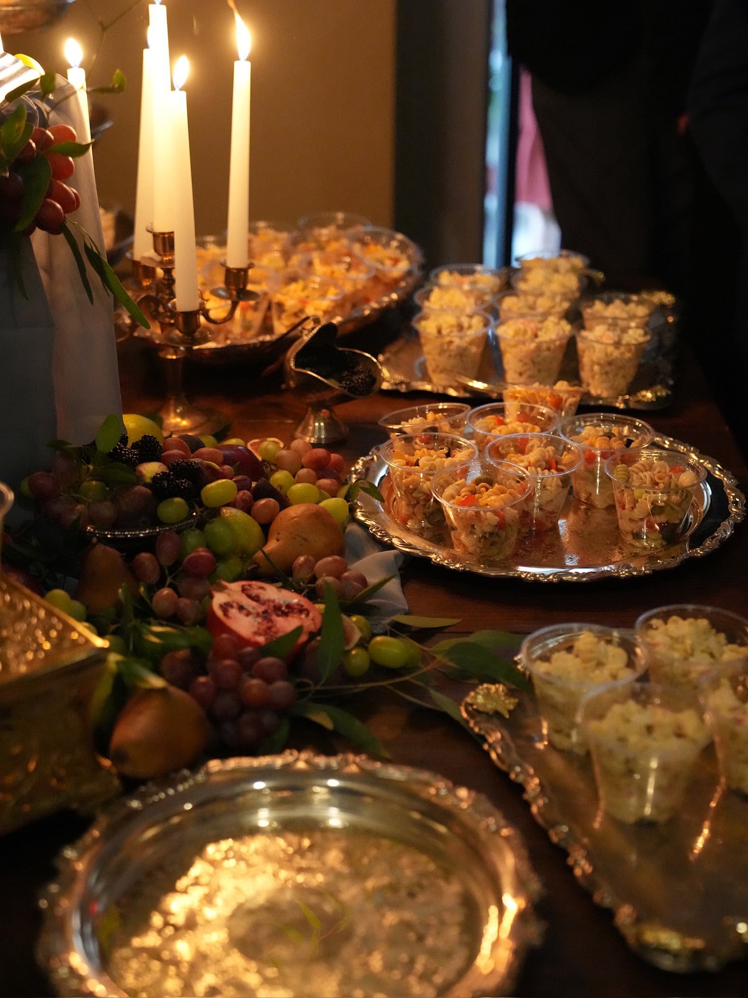 A table decorated for a celebration with an ornate candelabrum, fresh fruit including grapes, pears, and a pomegranate, and an assortment of colorful snacks in small cups, illuminated by candlelight.