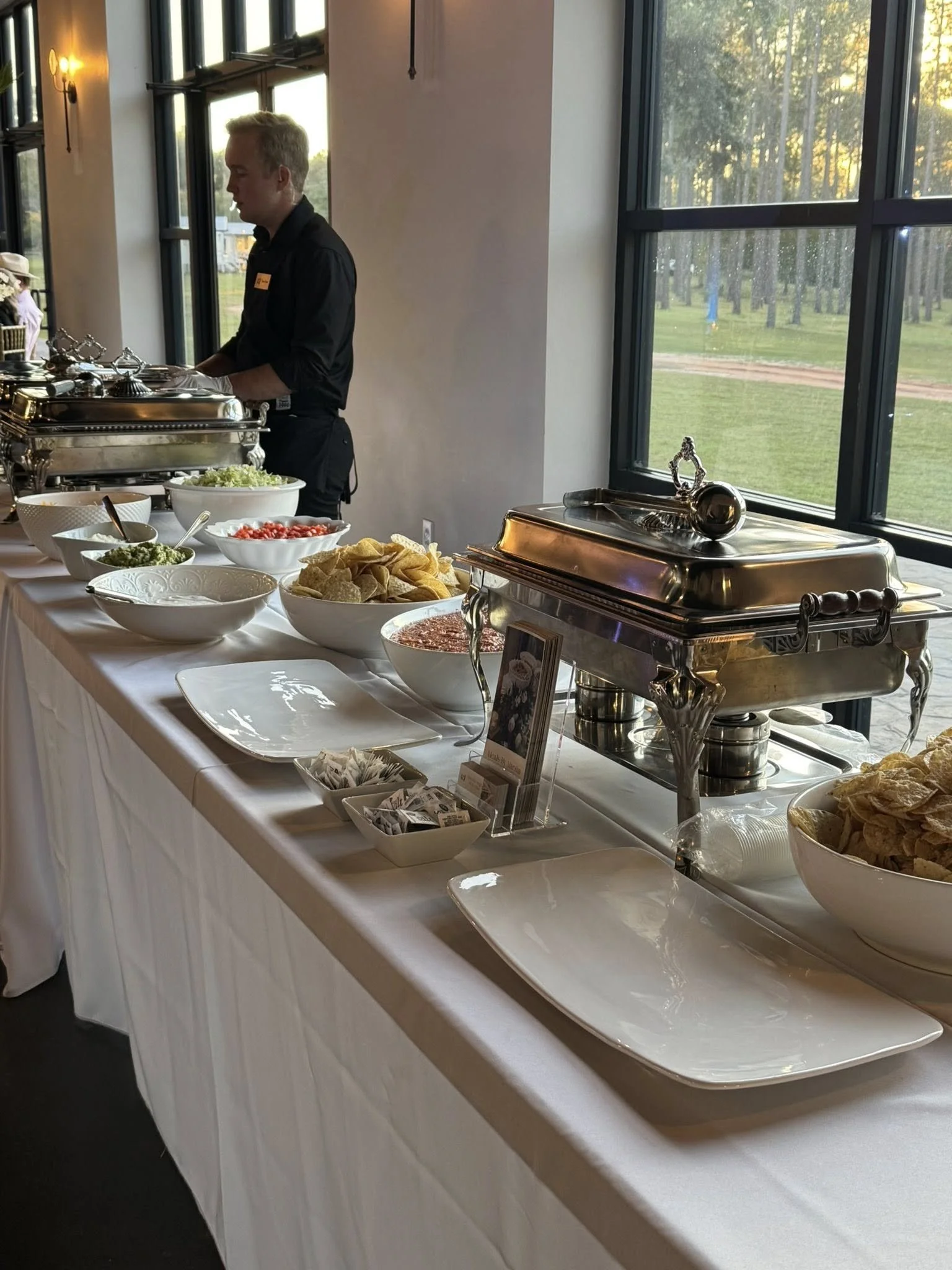 Buffet table with chafing dishes, bowls of chips, salsa, salad, and condiments, set up near large windows with a view of a golf course or park.