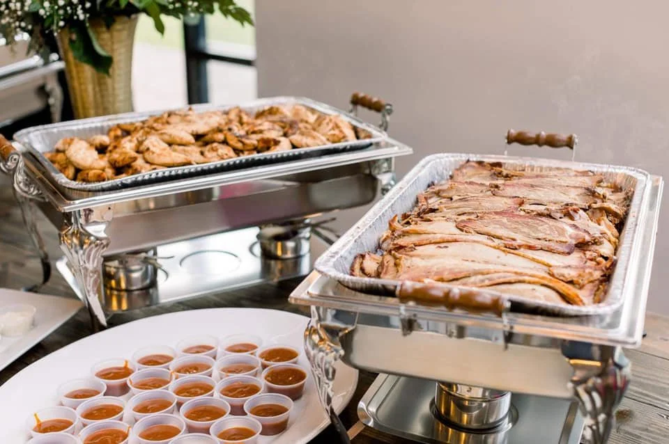 Chafing dishes with roasted chicken and sliced beef, along with small cups of barbecue sauce on a table at a buffet.