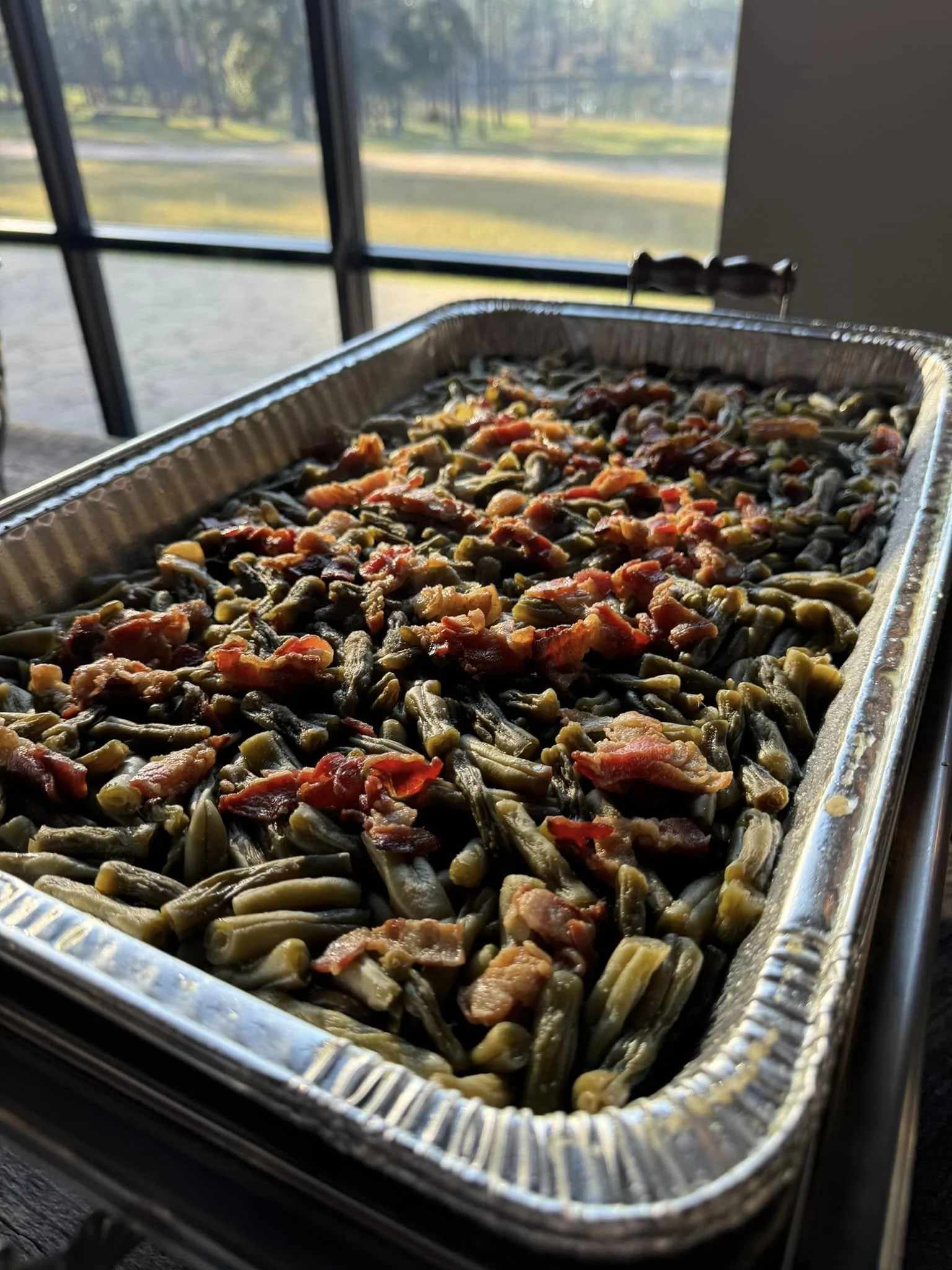 Baked green beans with bits of bacon in a foil tray on a table near a window showing a grassy outdoor area.