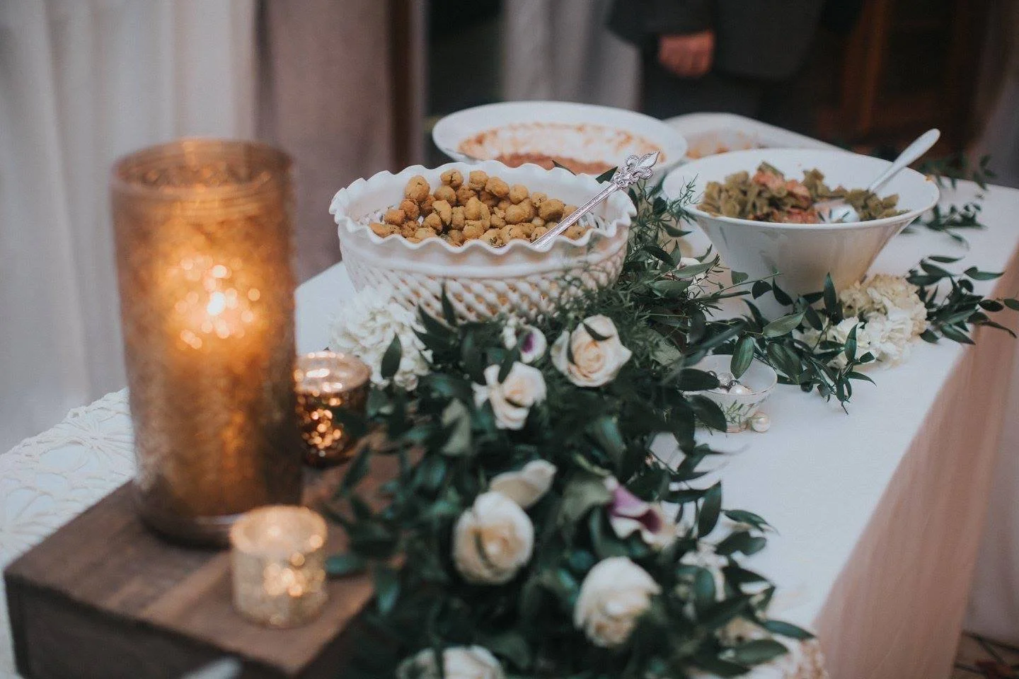 A table decorated with white roses and green foliage, featuring a large gold candle, a small candle, and a bowl of fried snacks. There are also bowls of salad and other dishes in the background.