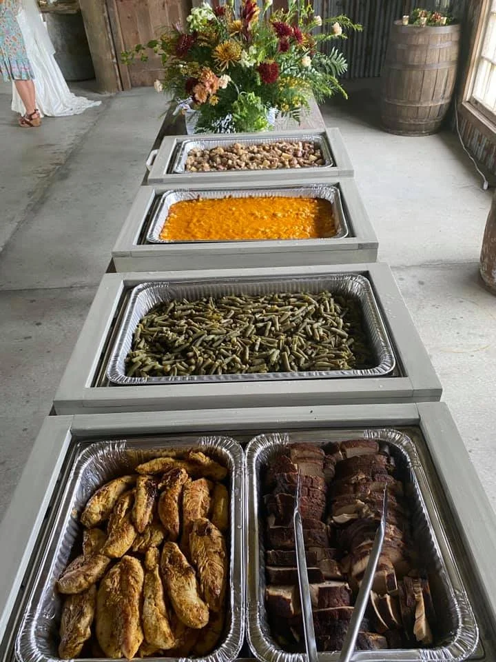 Buffet table with chicken wings, sliced beef, green beans, casserole, beans, and mashed potatoes with gravy, with a floral arrangement in the background.