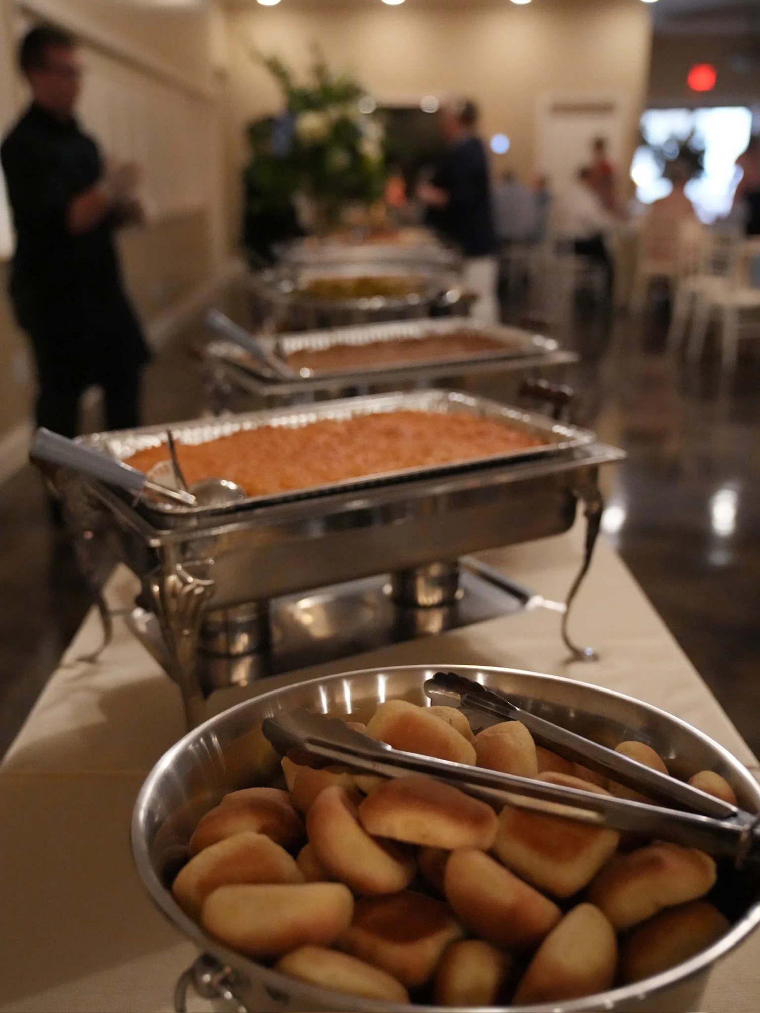 Buffet table with chafing dishes of hot food and a bowl of bread rolls in the foreground at a restaurant event.