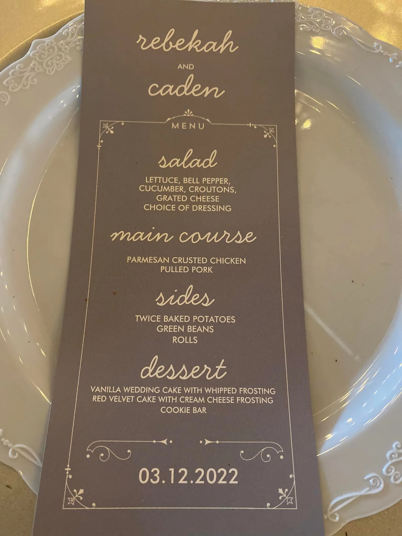 Wedding menu card with gold decorative border placed on a white plate with ornate edges. The menu includes salad, main course, sides, and dessert, with the date 03.12.2022 at the bottom.