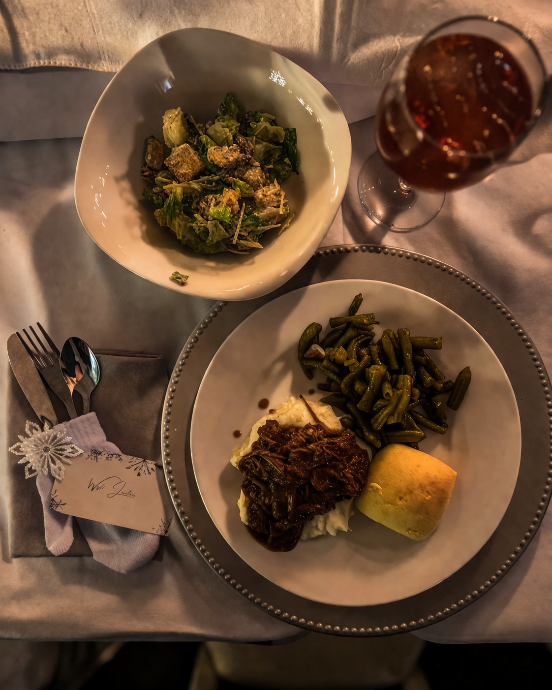 A dining table setting with a plate of mashed potatoes topped with beef stew, green beans, and an apple, a bowl of mixed greens salad, a glass of cola, and a napkin with a fork, knife, and spoon.