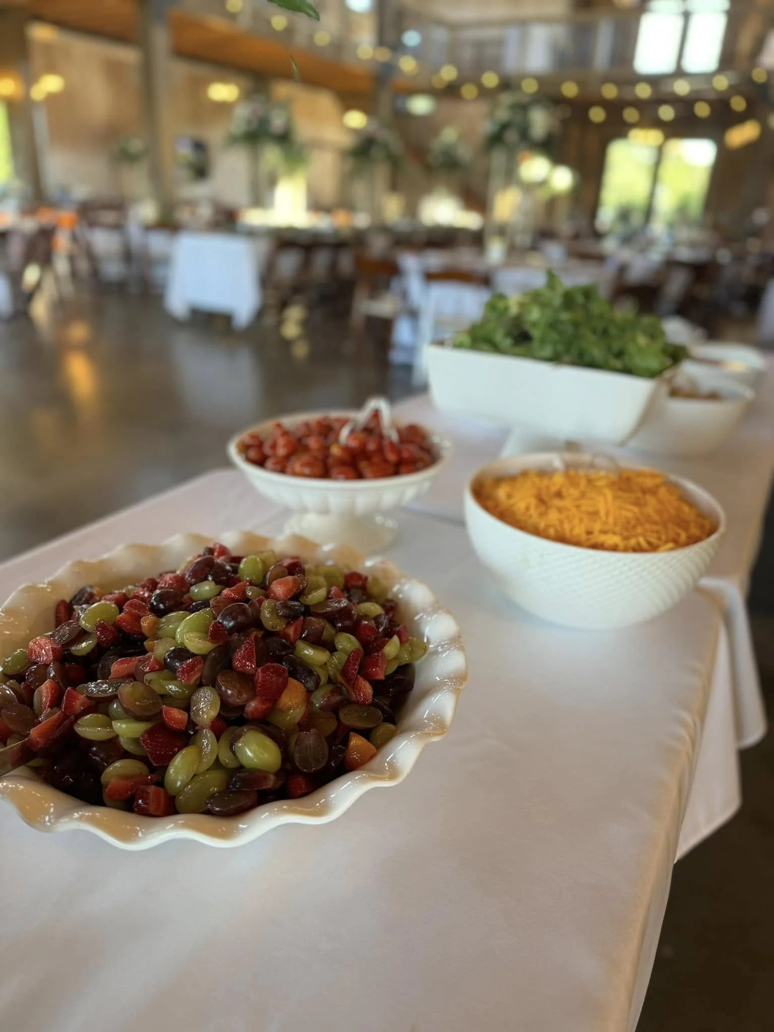 A table with bowls of grapes, cherry tomatoes, shredded cheese, and salad in a bright, decorated outdoor dining area.