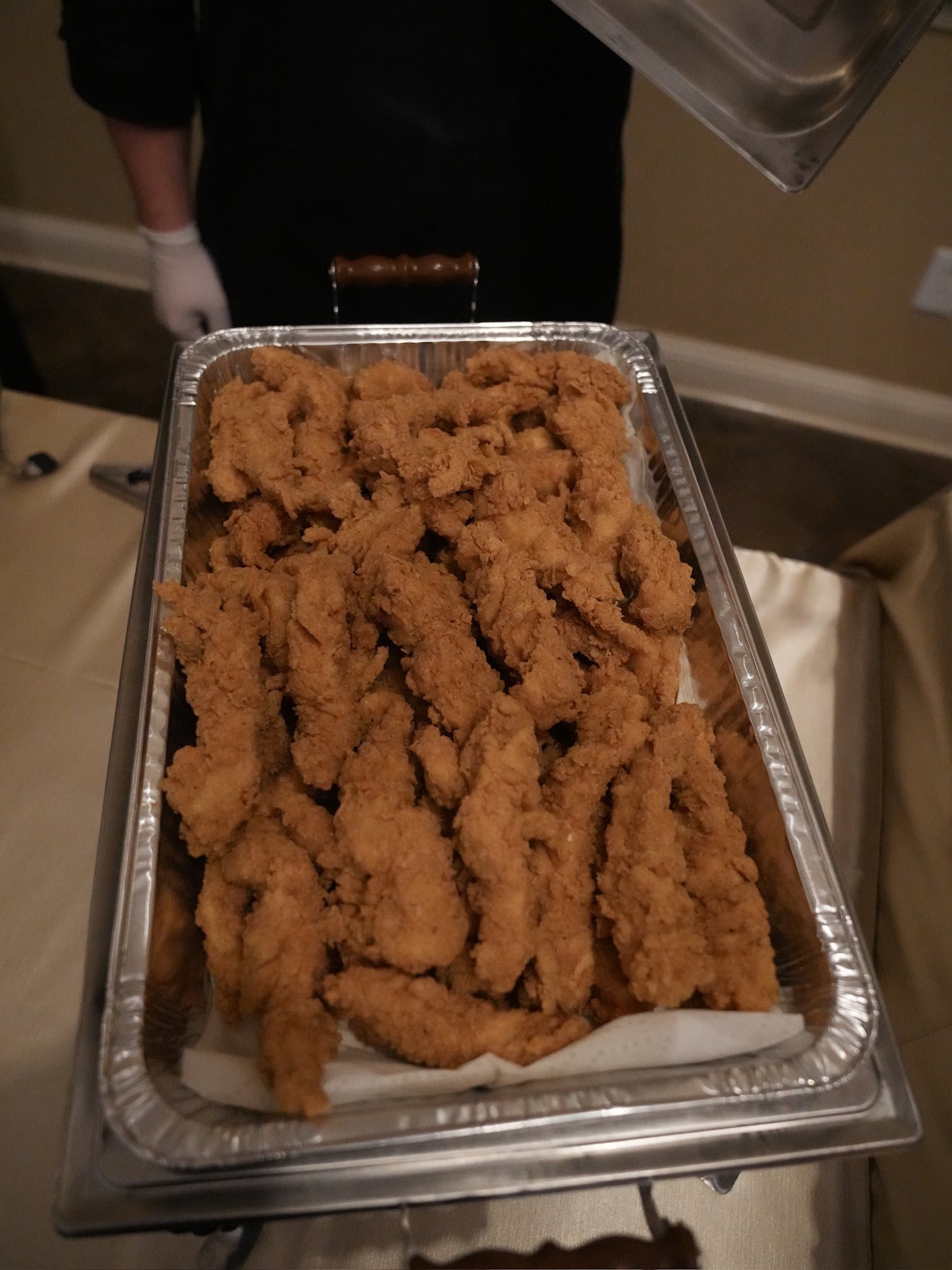 Tray of fried chicken tenders being held by a person in a dark uniform, with a person wearing white gloves behind, in an indoor setting.
