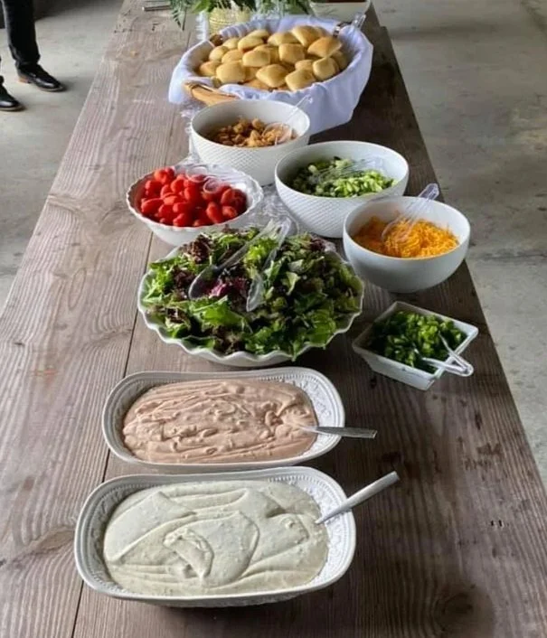 A wooden table with bowls of sandwiches, chopped vegetables, cherry tomatoes, salad, shredded cheese, and two types of dips or dressings.