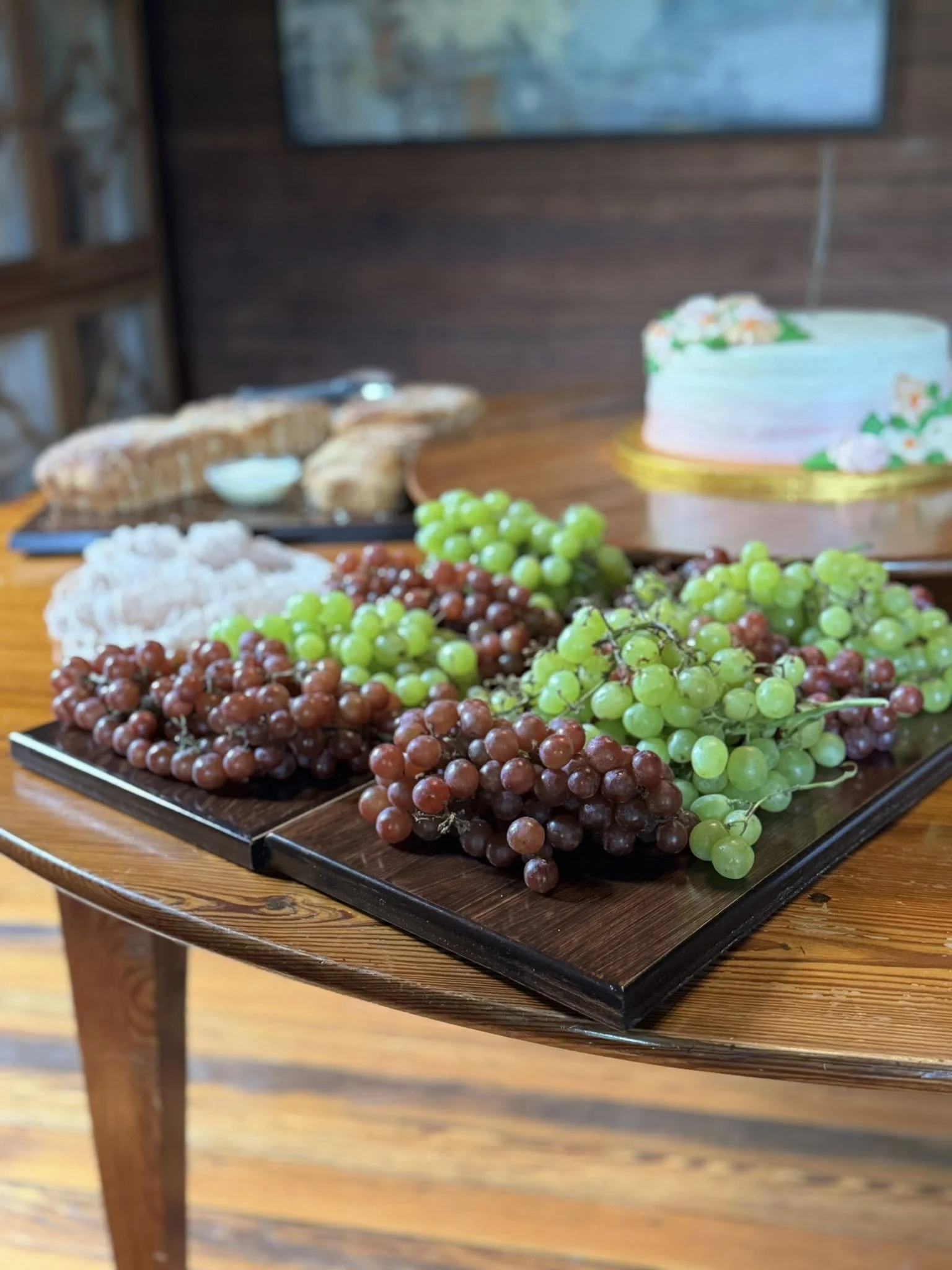 Grapes and children’s food on a black tray on a wooden table, with a birthday cake and bread in the background.
