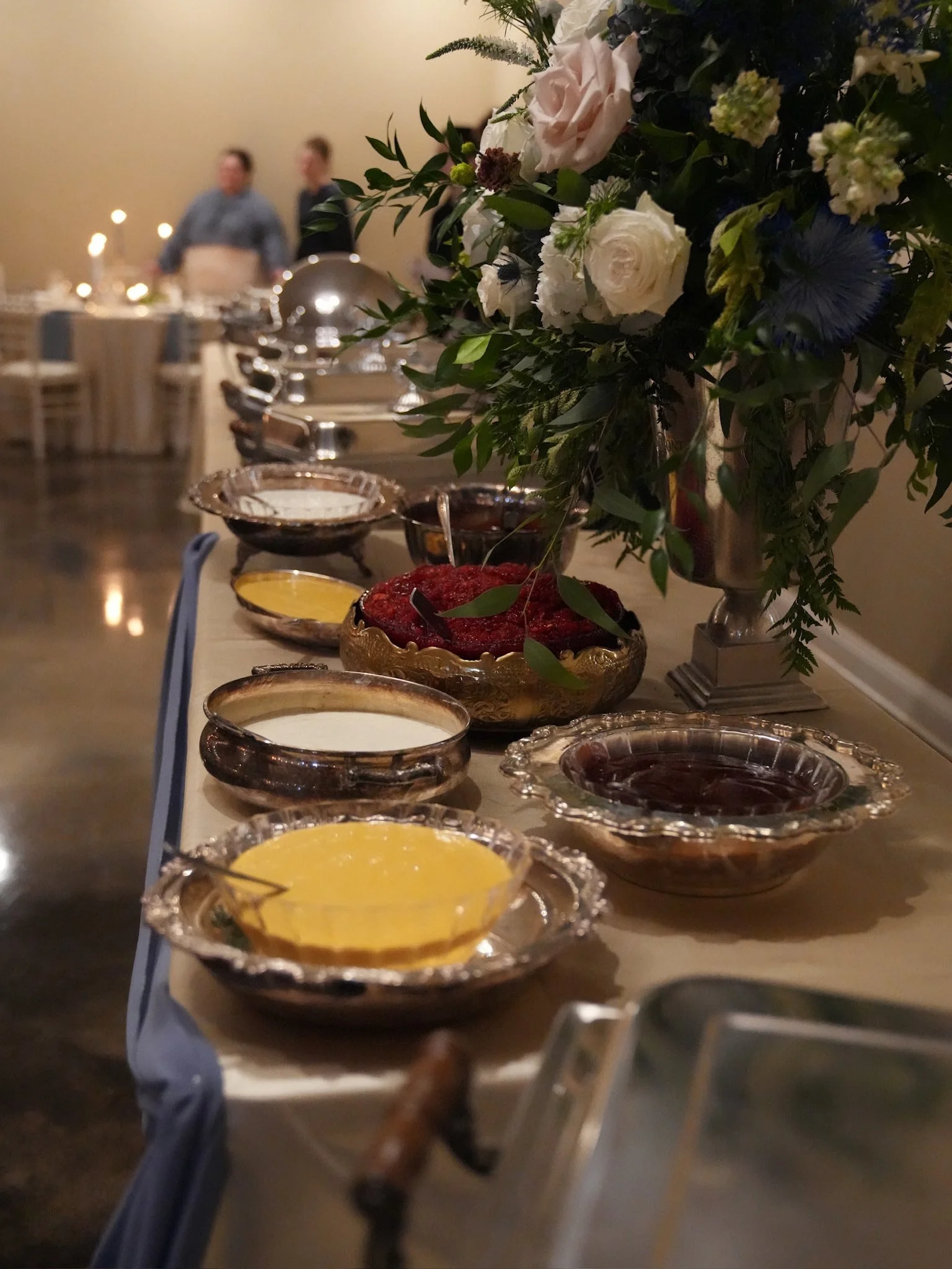 A buffet table with various sauces and dishes, decorated with a tall floral arrangement of white and pink roses, and other greenery. In the background, people are standing and talking in a warmly lit room setup for a formal event or celebration.