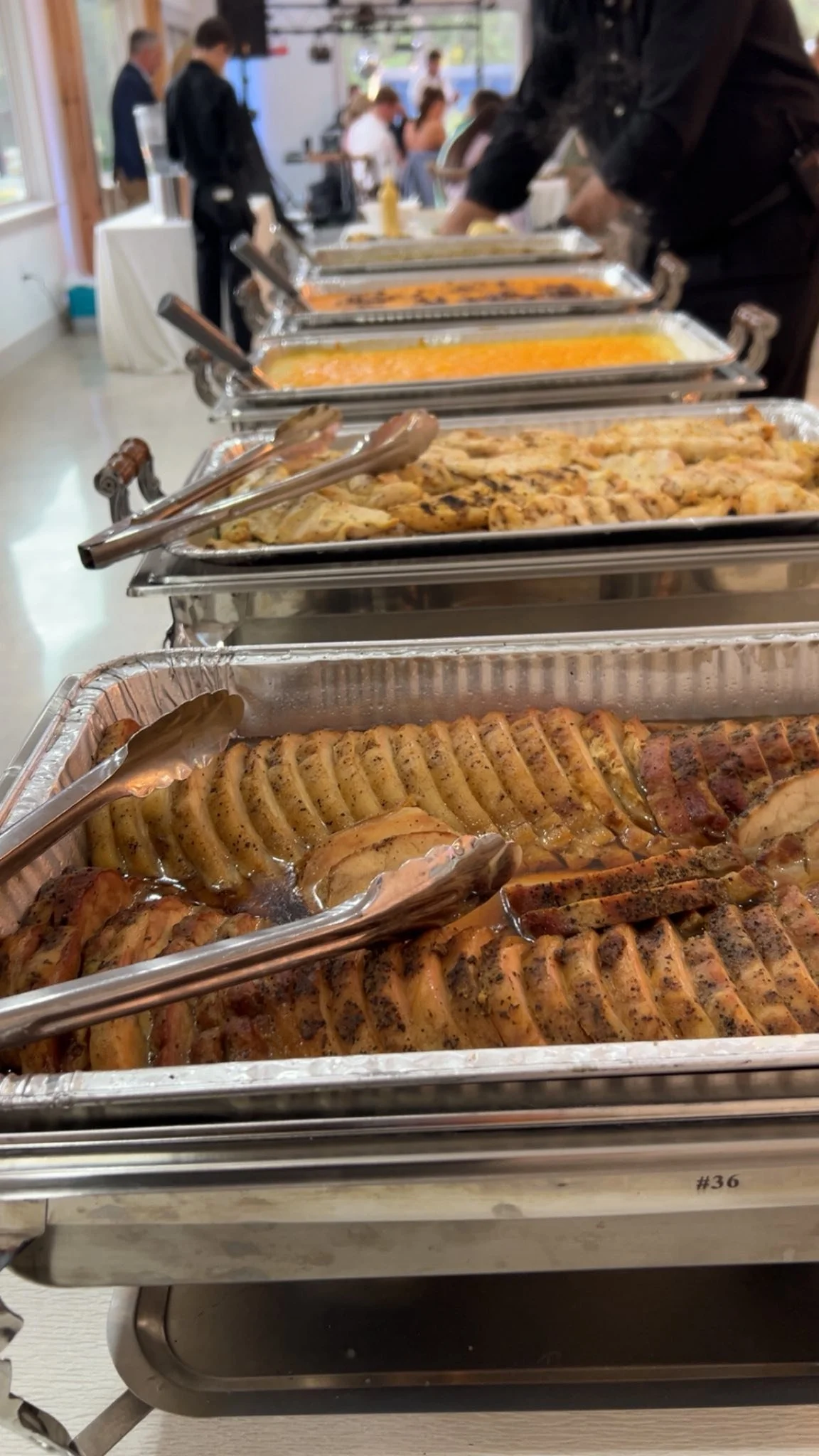 Buffet table with trays of grilled meats, roasted vegetables, and casseroles at a gathering or event.