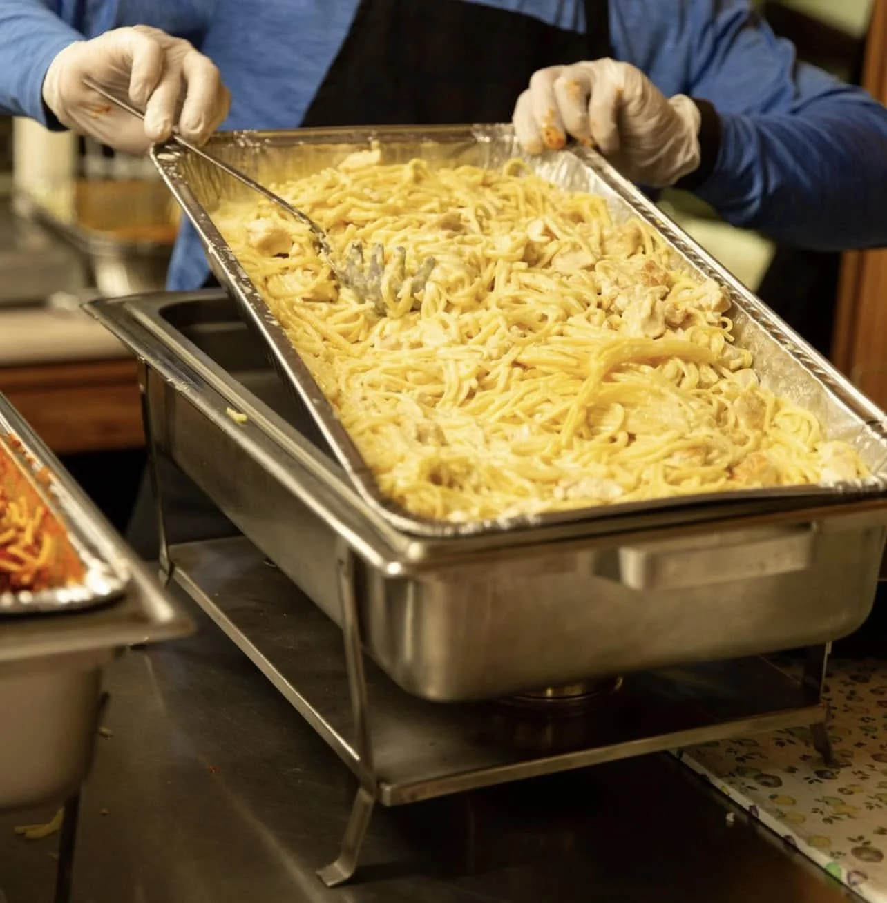 Person serving a large tray of creamy pasta with chicken, using tongs, wearing gloves, in a buffet setting.