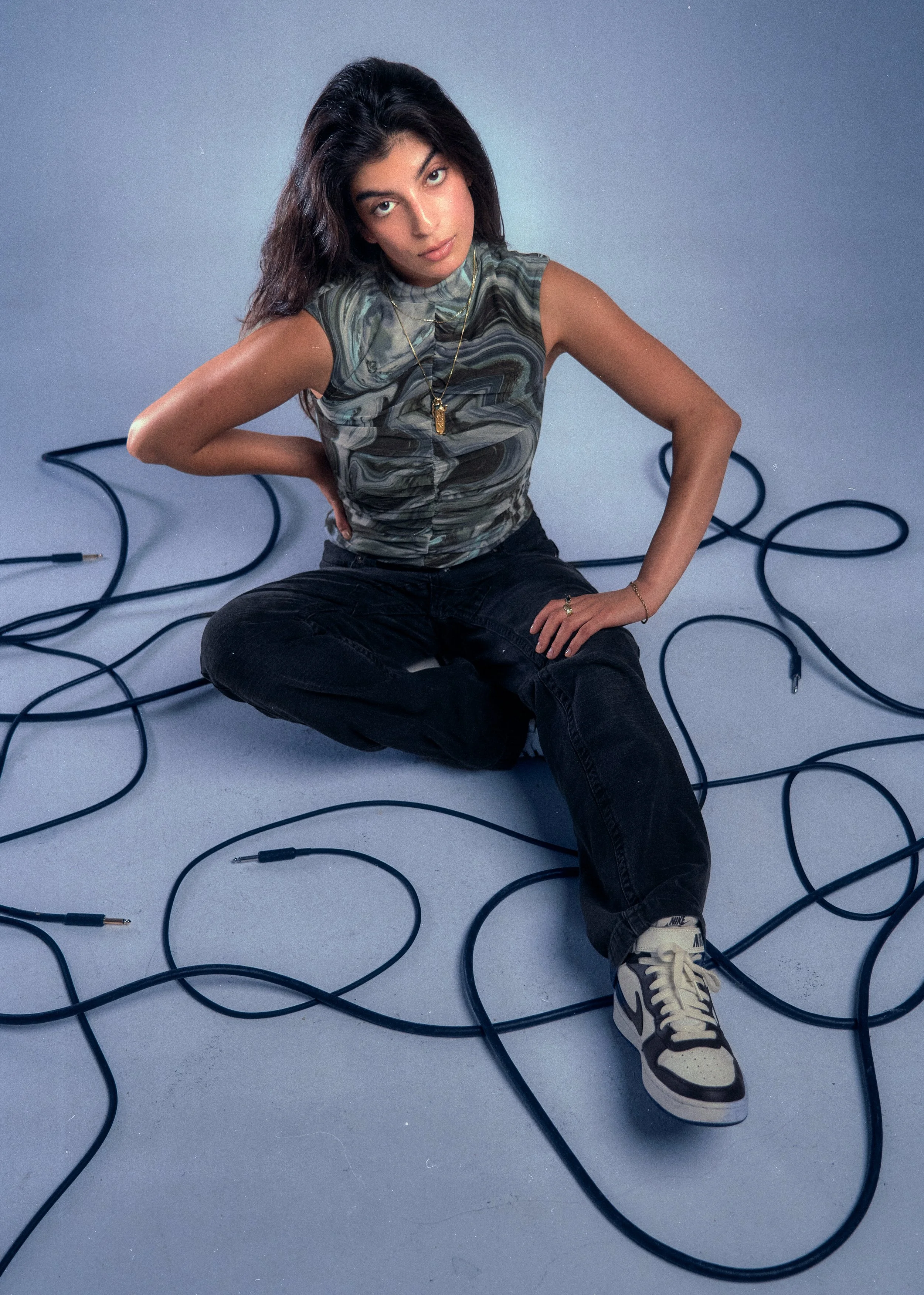 A woman sitting on the floor surrounded by tangled black electrical cords, wearing a patterned sleeveless top, black pants, and Nike sneakers, with her hand on her hip and looking at the camera.