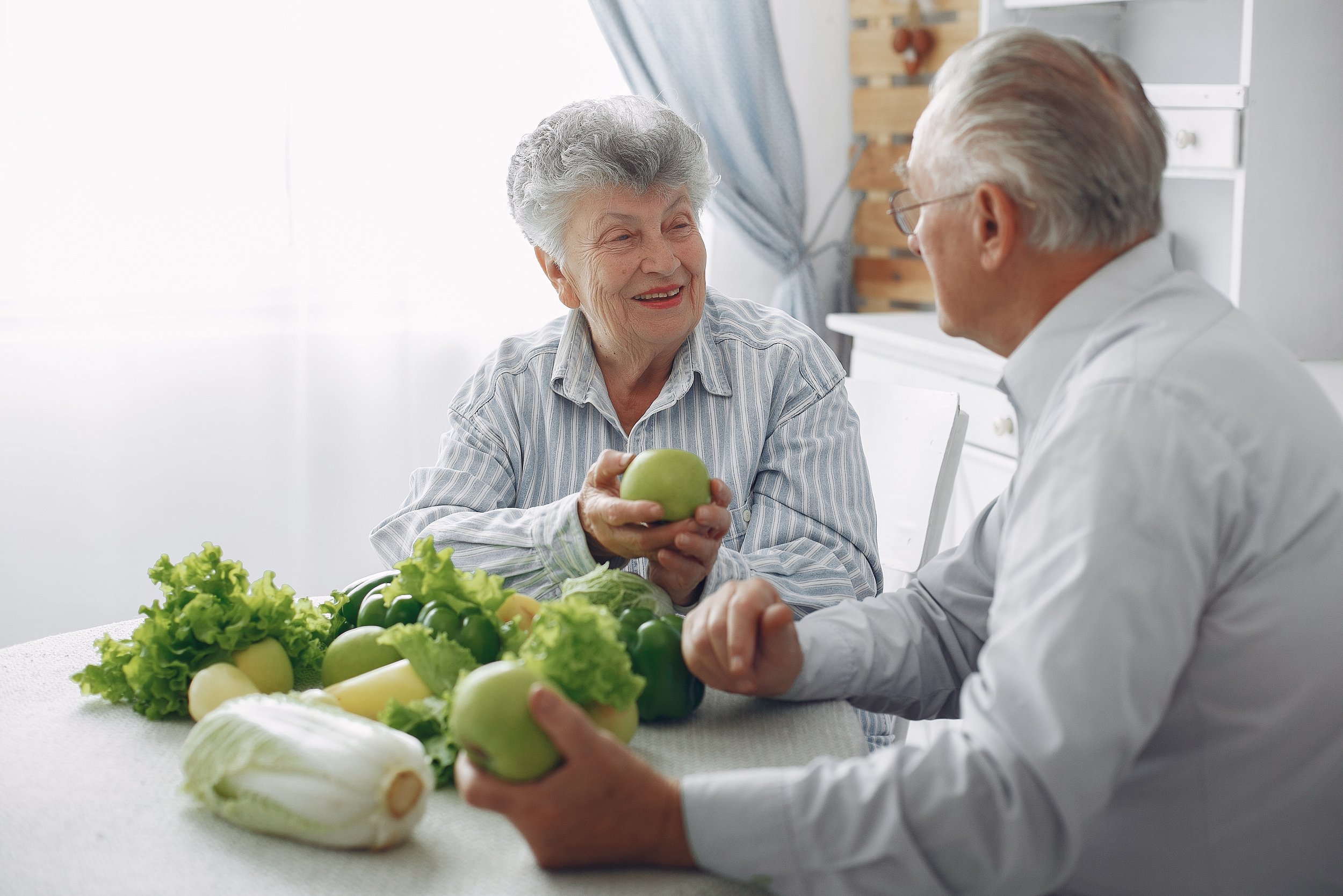 An elderly woman and a middle-aged man are sitting at a table with green apples and various greens. The woman is smiling and holding an apple, while the man hands an apple to her. They are indoors in a bright room with kitchen cabinets in the background.