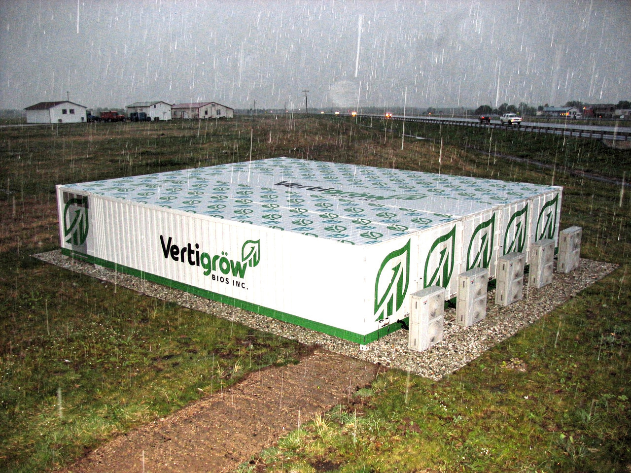A large container labeled 'Vertigrow BIOS INC.' is outdoors in the rain, with a gravel base and surrounded by grass. There are several air conditioning units attached to the side of the container. The background features a rural area with houses, power lines, and a highway.