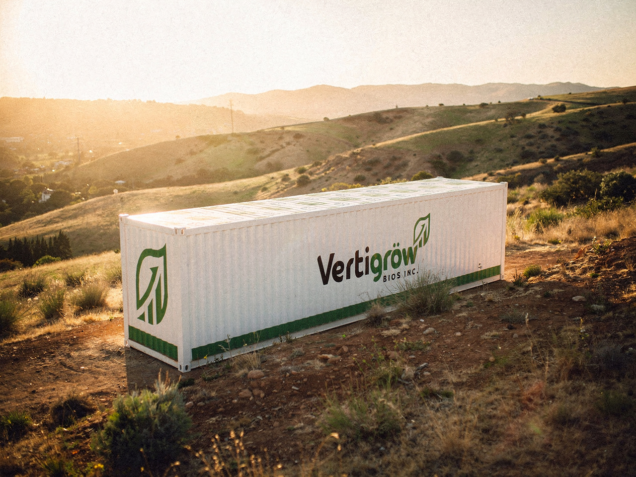 A white shipping container with green logo and text reading 'Vertigrow BIOS INC' on a hilltop with grassy, rocky terrain, open landscape, and hills in the background during sunset.