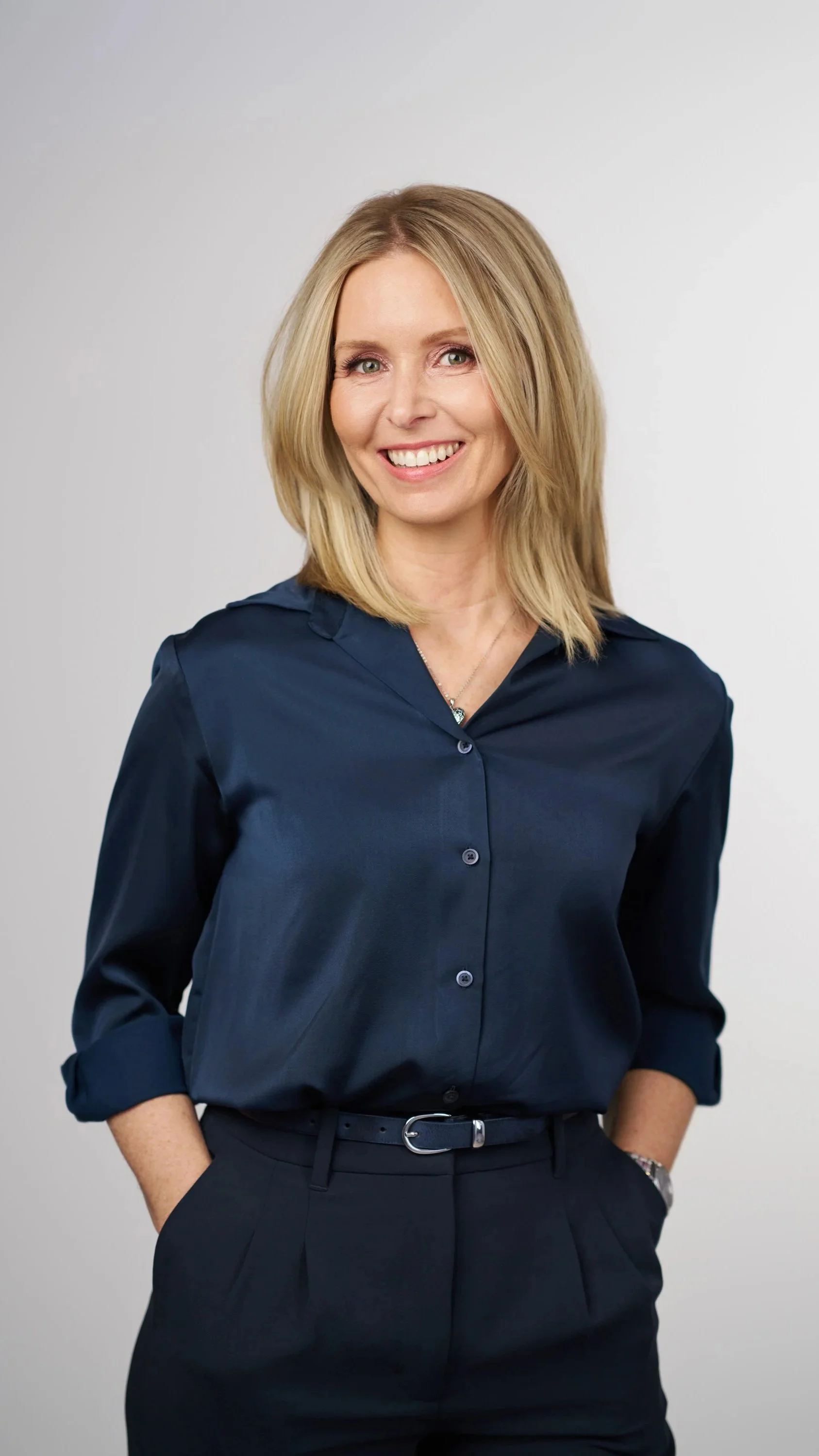 A woman with shoulder-length blonde hair is smiling and looking at the camera. She's wearing a dark blue button-up shirt with the sleeves rolled up and dark pants with a belt. Her hands are in her pockets, and she is standing against a plain gray background.