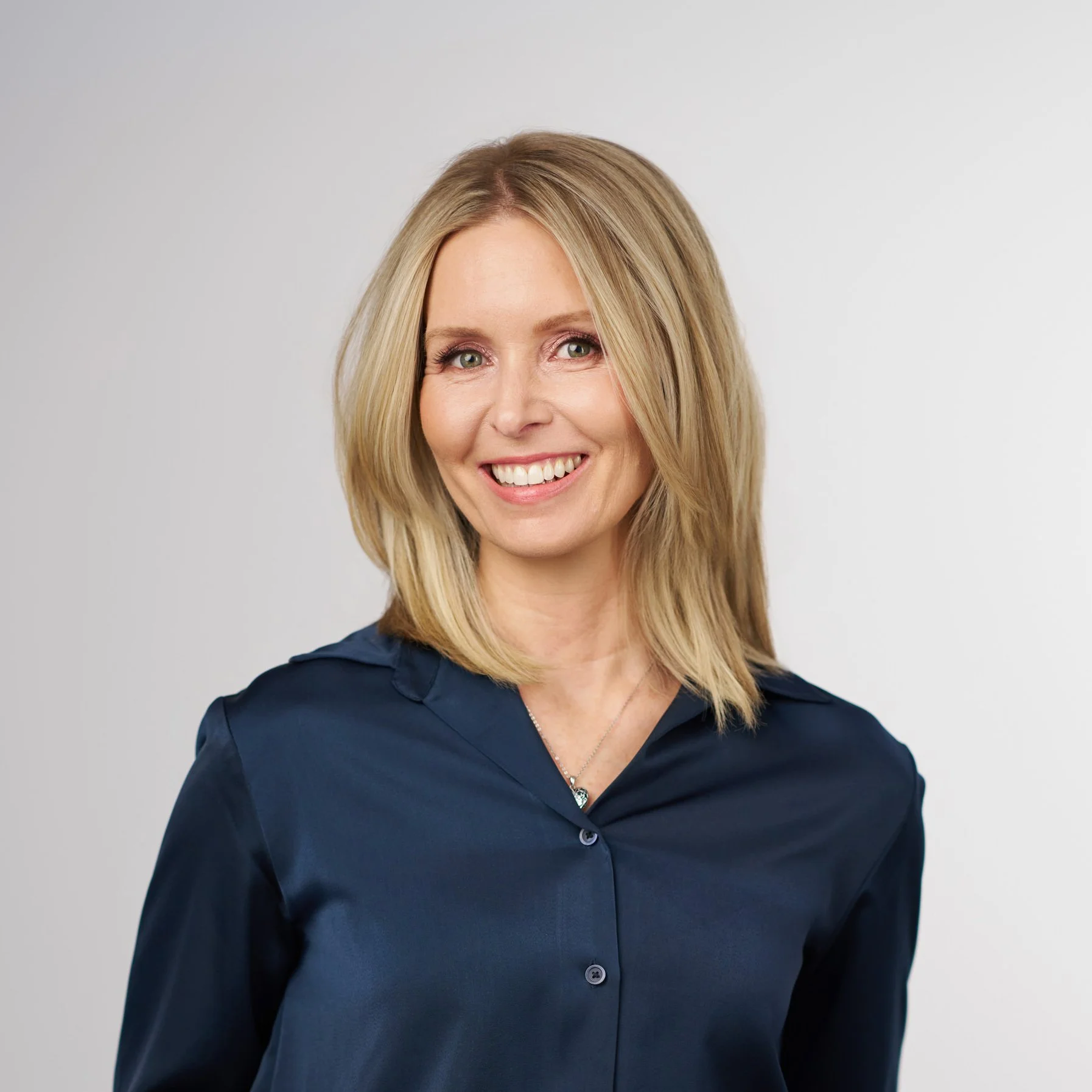 A smiling woman with shoulder-length blonde hair, wearing a dark blue button-up shirt, standing against a plain light gray background.