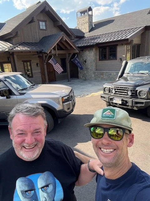 Two men smiling in front of a house with two parked trucks, American flag, and a DNC flag, on a sunny day.