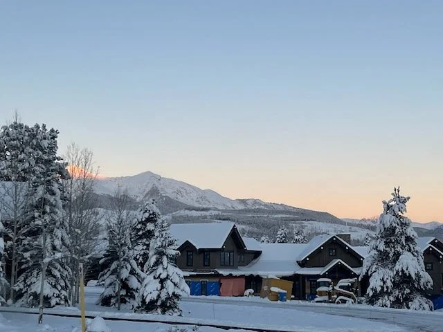 Snow-covered houses and trees in a winter landscape with mountains in the background during sunset.