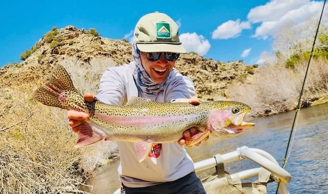Person wearing sunglasses, a cap, and a long-sleeve shirt holding a large rainbow trout near a riverbank with rocky hills and blue sky in the background.