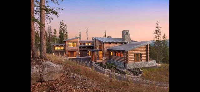 Large modern log cabin with multiple levels and large windows, situated on a hillside surrounded by trees at sunset.