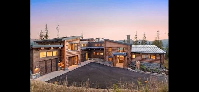 A modern log cabin-style house with wooden exterior and large windows, situated in a natural setting with trees and a dirt driveway.